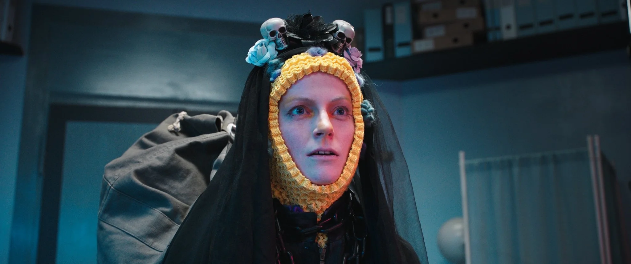 Frau mit Frida Kahlo-inspiriertem Make-up und einem Kopfband mit kleinen Totenkopf- und Blumenmotiv, trägt schwarze Kleidung und einen Rucksack, in einem Raum mit blauer Beleuchtung.