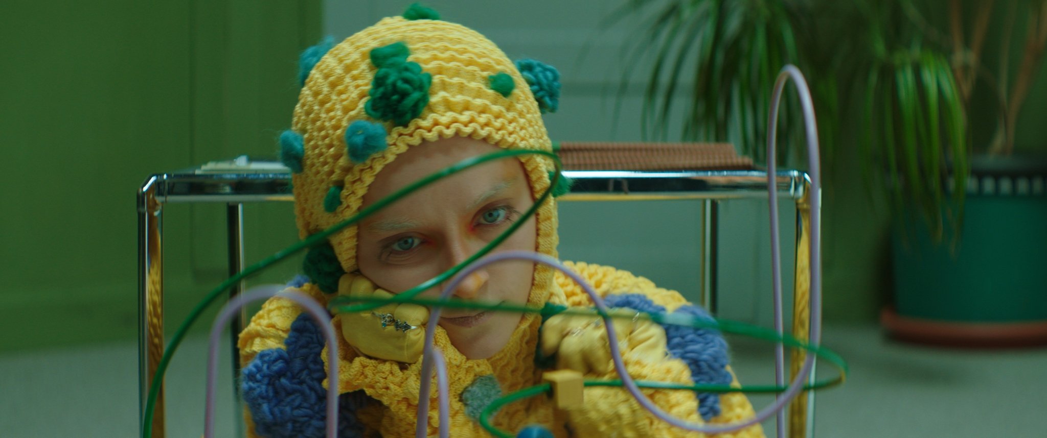 Nahaufnahme einer Person mit blauen Augen, die ein knallgelbes, gestricktes Haube trägt, umgeben von bunten Drähten und elektronischen Komponenten, vermutlich in einem futuristischen oder künstlerischen Setting.