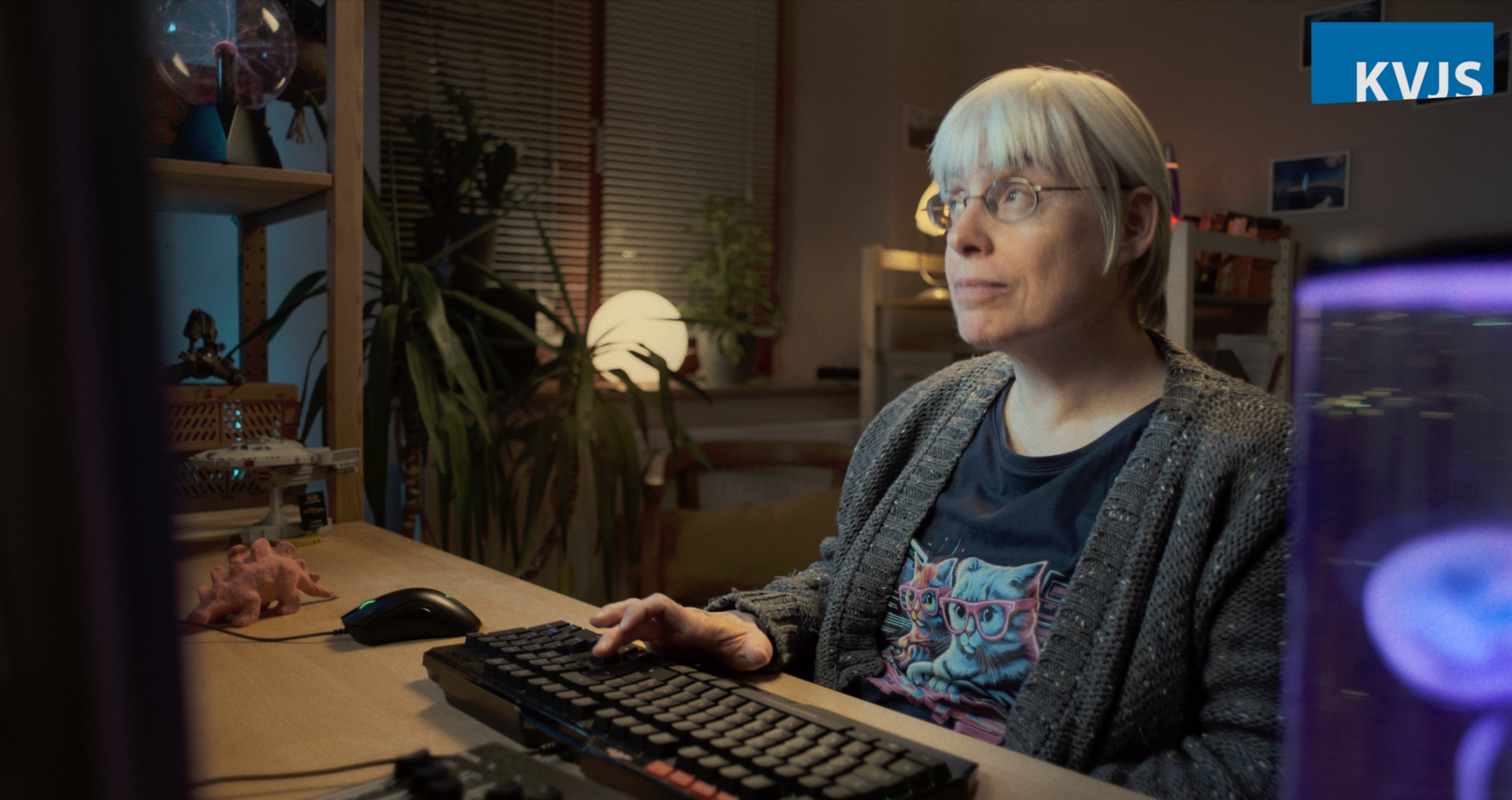 Ein älterer Mann mit Brille sitzt vor einem Computerbildschirm in einem Raum mit Pflanzen und Bücherregalen, im Hintergrund sind Fenster mit geschlossenen Jalousien.