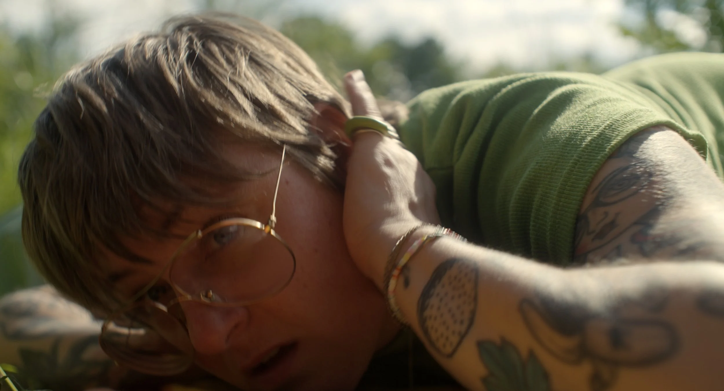 Nahaufnahme einer Person mit Brille, die auf dem Boden liegt, bei sonnigem Wetter im Freien, mit grünen Bäumen im Hintergrund.