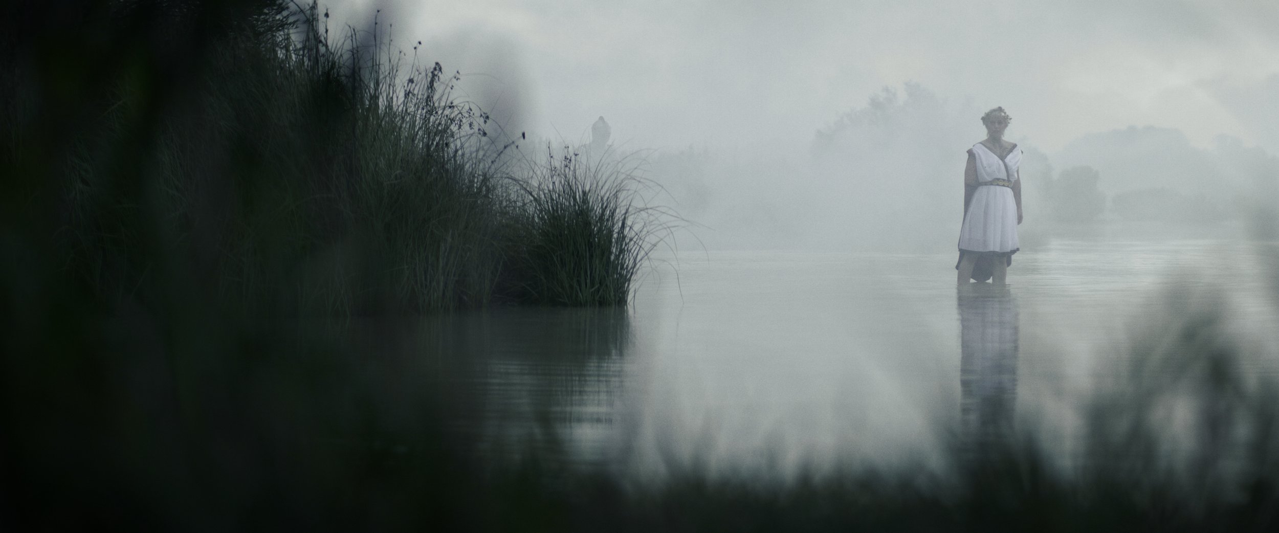 Eine Frau steht knietief im Wasser in einer nebligen, naturbelassenen Umgebung, mit Gräsern und Bäumen im Hintergrund.