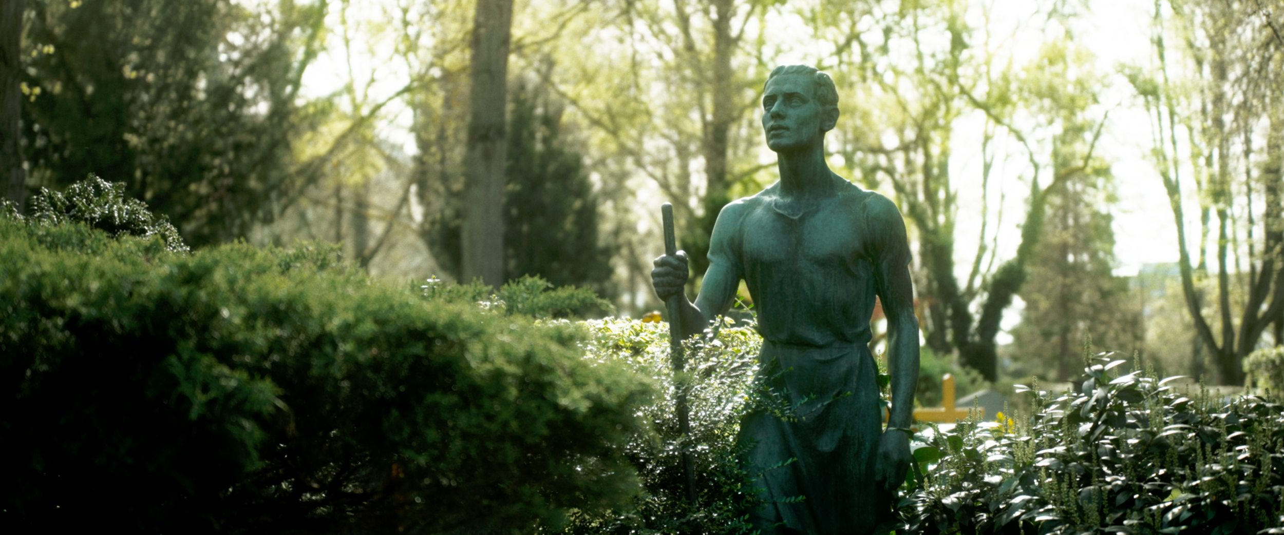 Bronzene Skulptur eines Mannes in einem Park, umgeben von Büschen und Bäumen im Sonnenlicht.