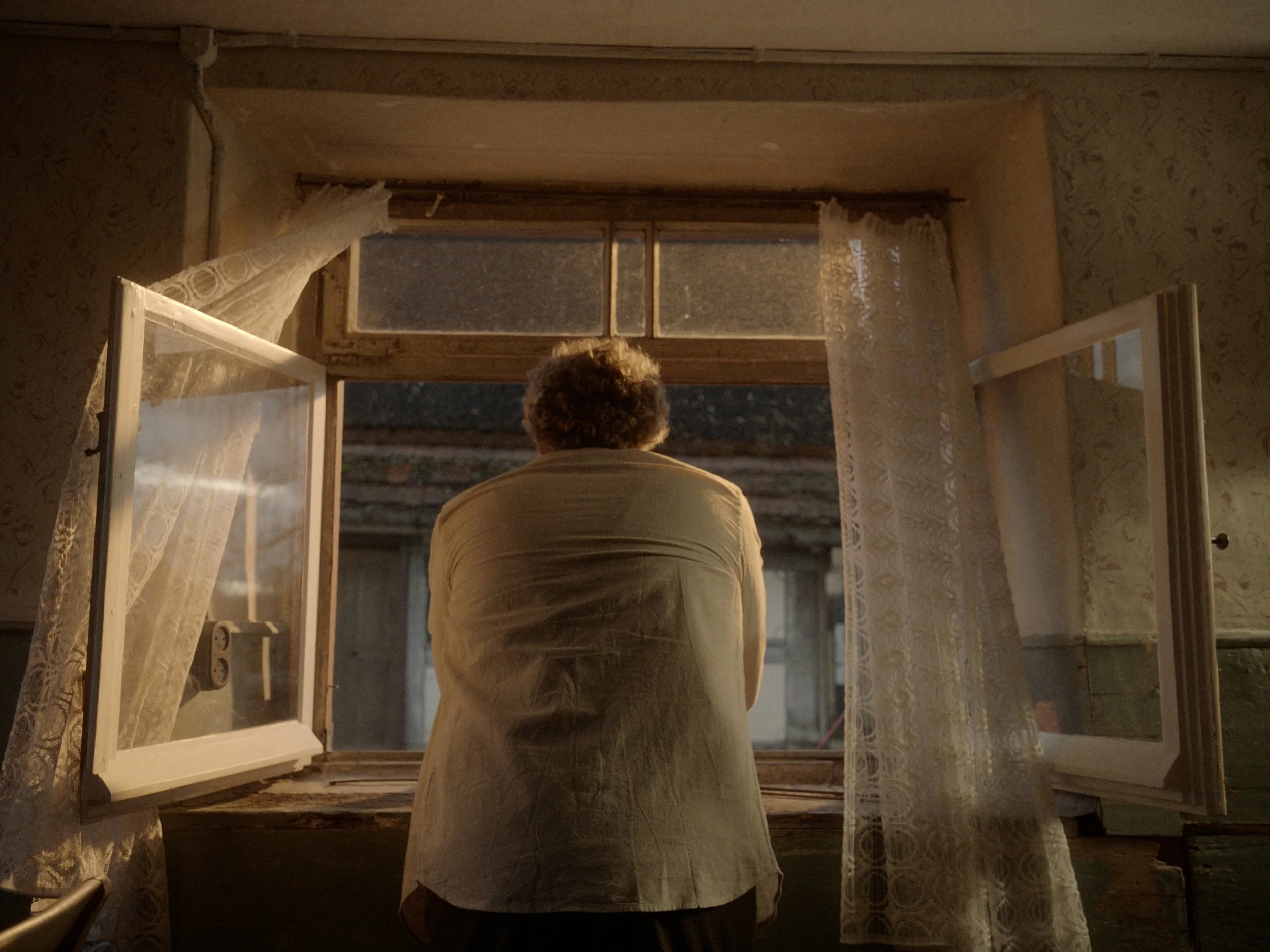 Ein älterer Mensch schaut aus einem offenen Fenster bei Dämmerung oder Nacht, im Raum hängen weiße Spitze-Vorhänge an den Fenstern.