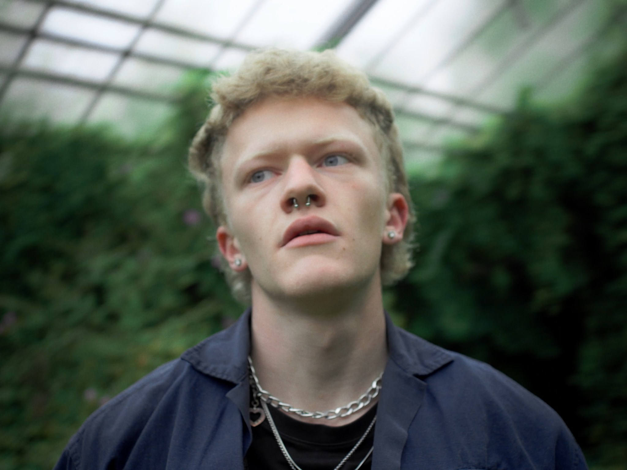 Nahaufnahme eines jungen Mannes mit kurzen, lockigen blonden Haaren, Piercings an der Nase und Ohr, trägt eine silberne Kette mit Anhänger und eine dunkle Jacke, im Hintergrund grünen Pflanzen in einem Gewächshaus.