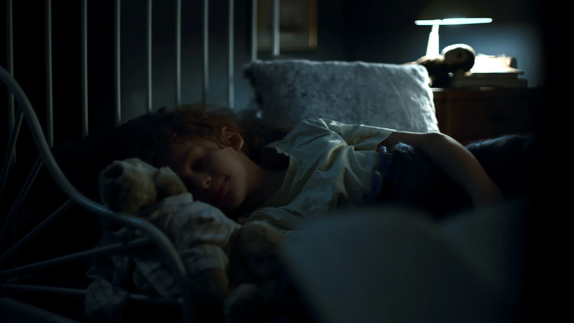 Ein Kind schläft friedlich in einem Bett, umgeben von dunkler Beleuchtung, mit einem Kuscheltier und einem Smartphone auf dem Nachttisch im Hintergrund.