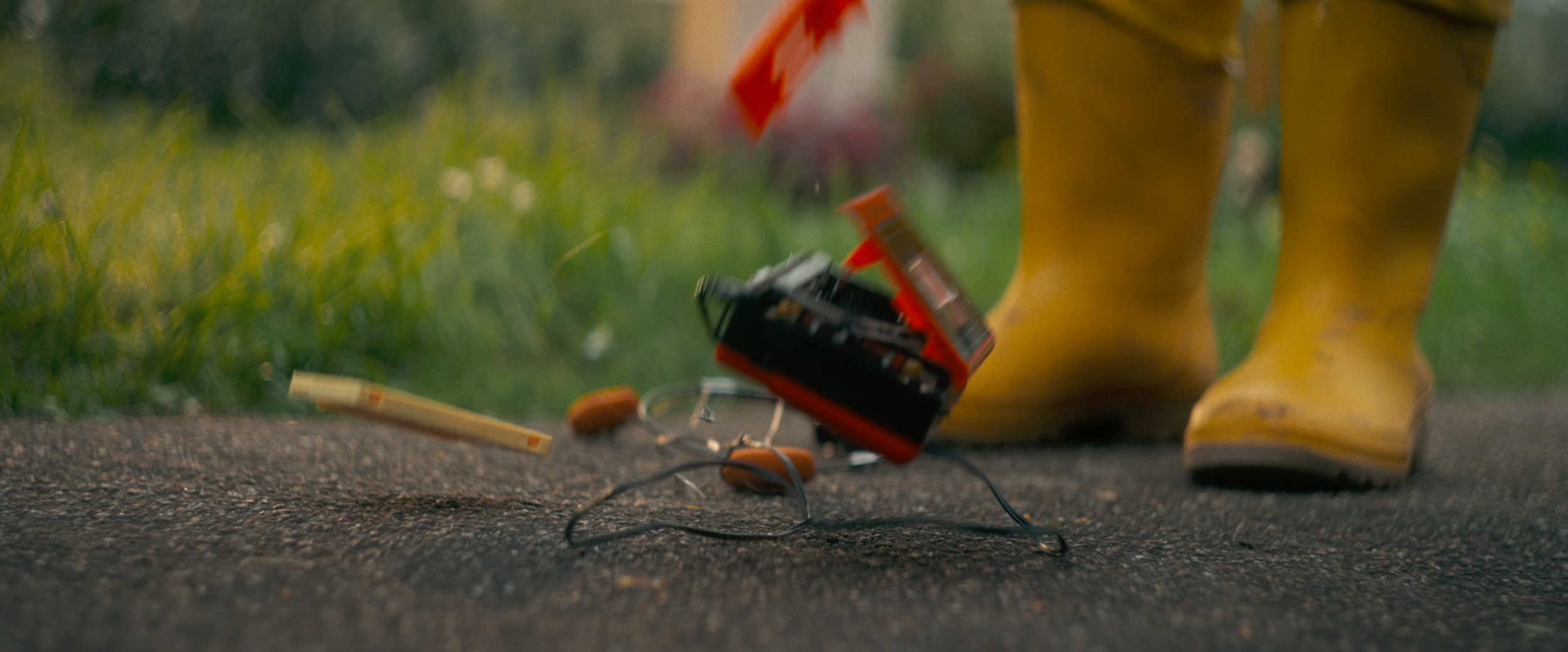 Ein Paar gelbe Gummistiefel steht auf Asphalt neben einem Schmutzrasen, mit verstreutem Werkzeug, darunter eine Batteriebox, Zündkerze, Zündkabel und eine kleine Holzleiste.