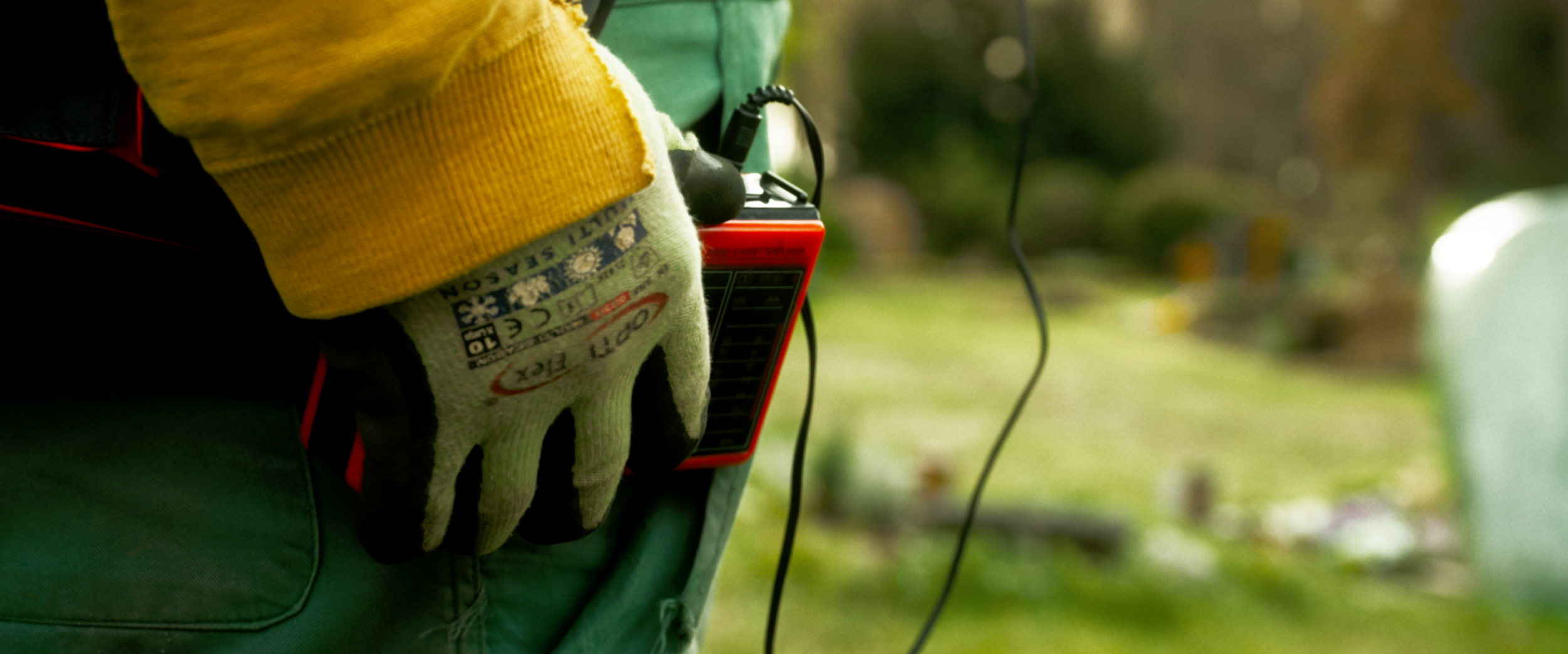 Eine Hand trägt eine graue Arbeitshandschuh und hält ein rotes Messgerät mit Kabel, die Person trägt eine gelbe Quasti, eine graue Jacke mit grüner Kleidung darunter, im Hintergrund ist eine grüne, outdoor Umgebung sichtbar.