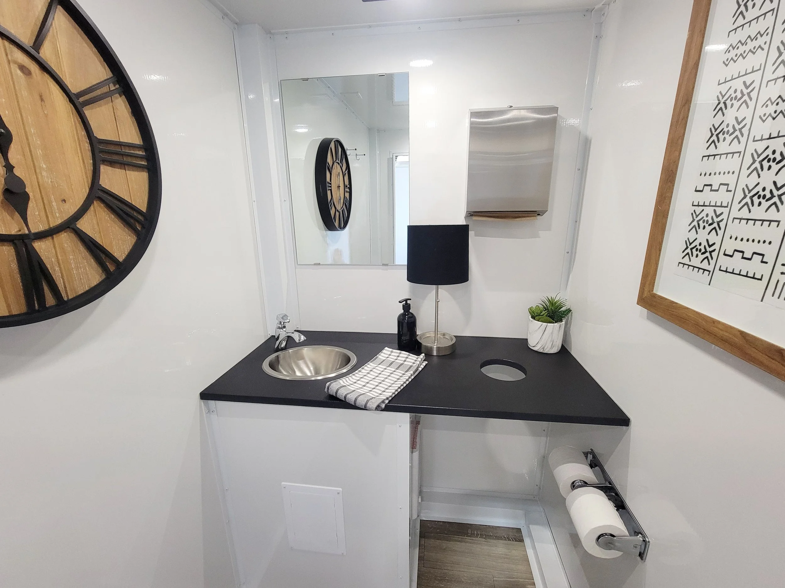 Small kitchenette with a black countertop, a stainless steel sink, a black lamp, a small potted plant, a soap dispenser, a mirror, a large clock, and framed wall art.
