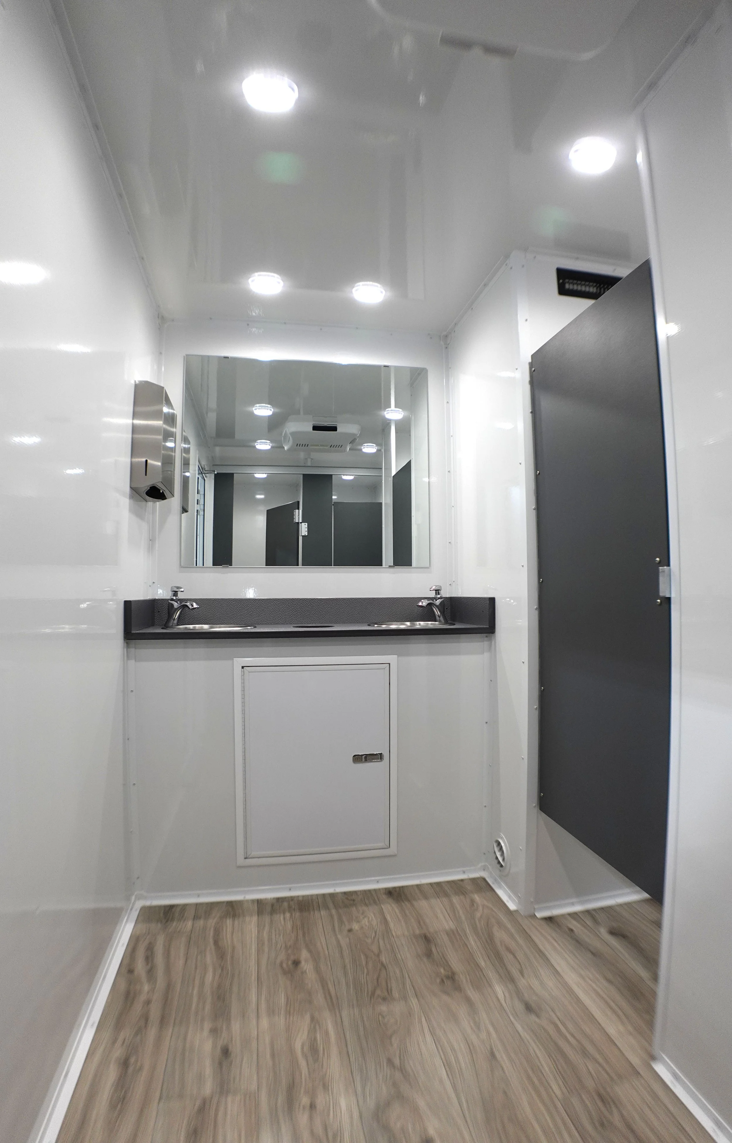 A small, clean bathroom with a wall-mounted mirror, two sinks, a lockable cabinet door underneath, and wood-patterned flooring.