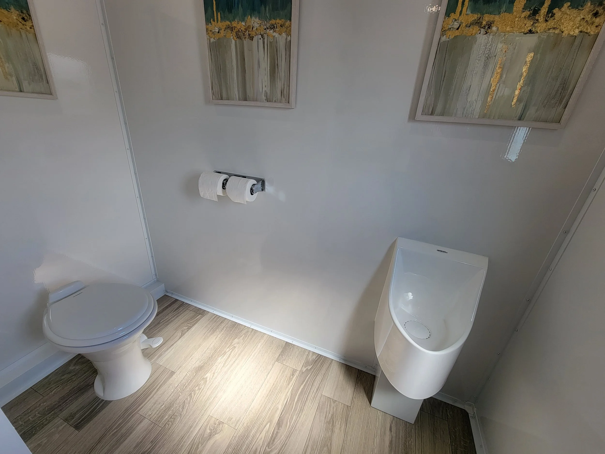 Modern bathroom with a toilet, a toilet paper holder with two rolls, a white urinal, and three framed abstract paintings on the wall.
