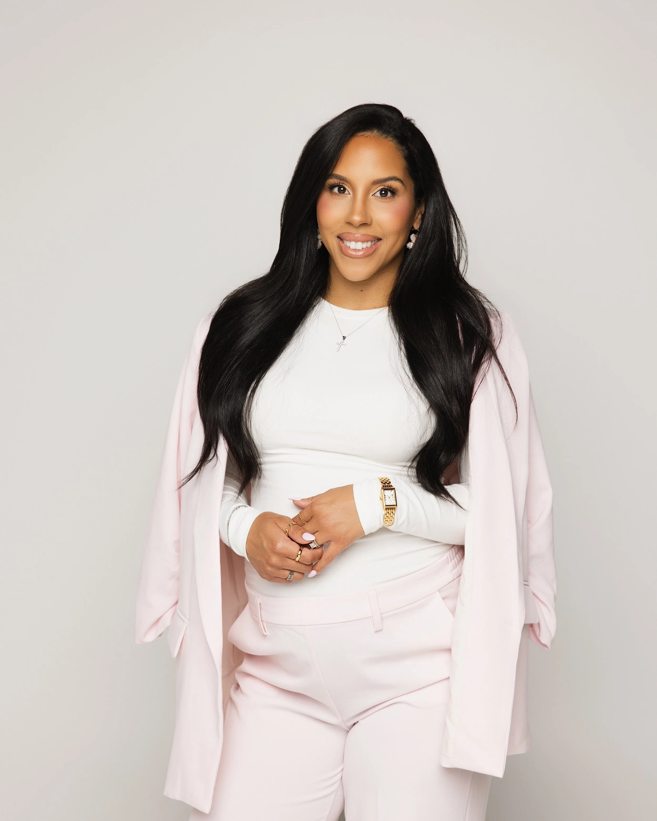 A woman dressed in a white top and pink suit with long black hair, jewelry, and a gold watch, standing against a plain light background.