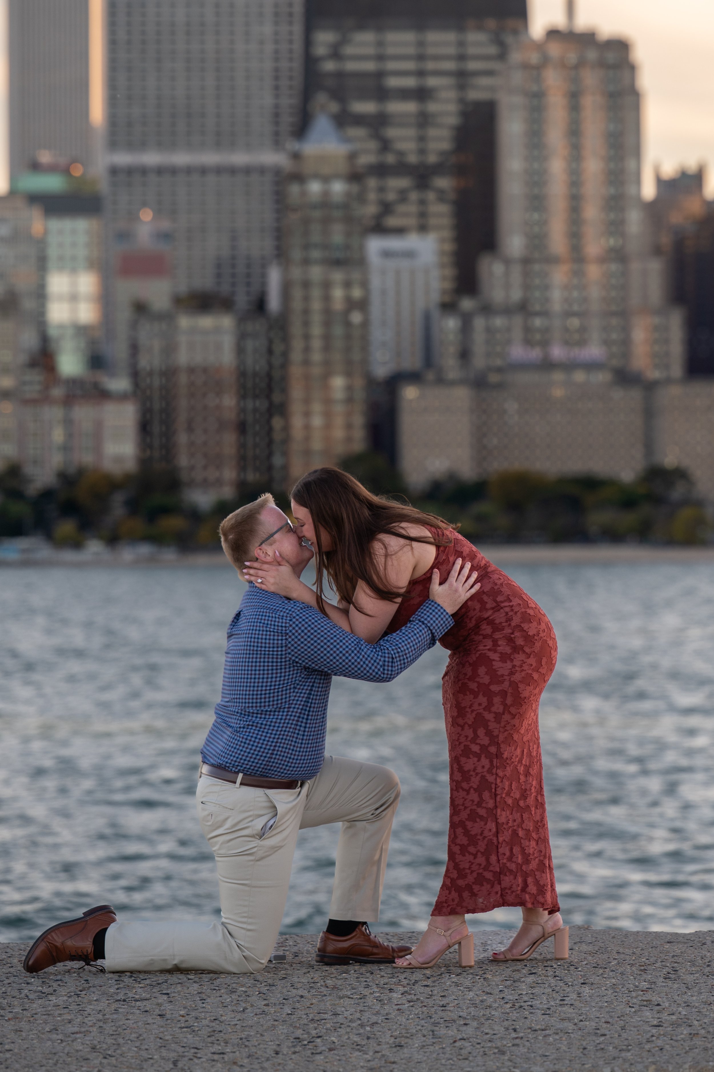 A couple is sharing a romantic moment by the water with the Chicago cityscape in the background, one person is kneeling and the other is leaning in for a kiss.