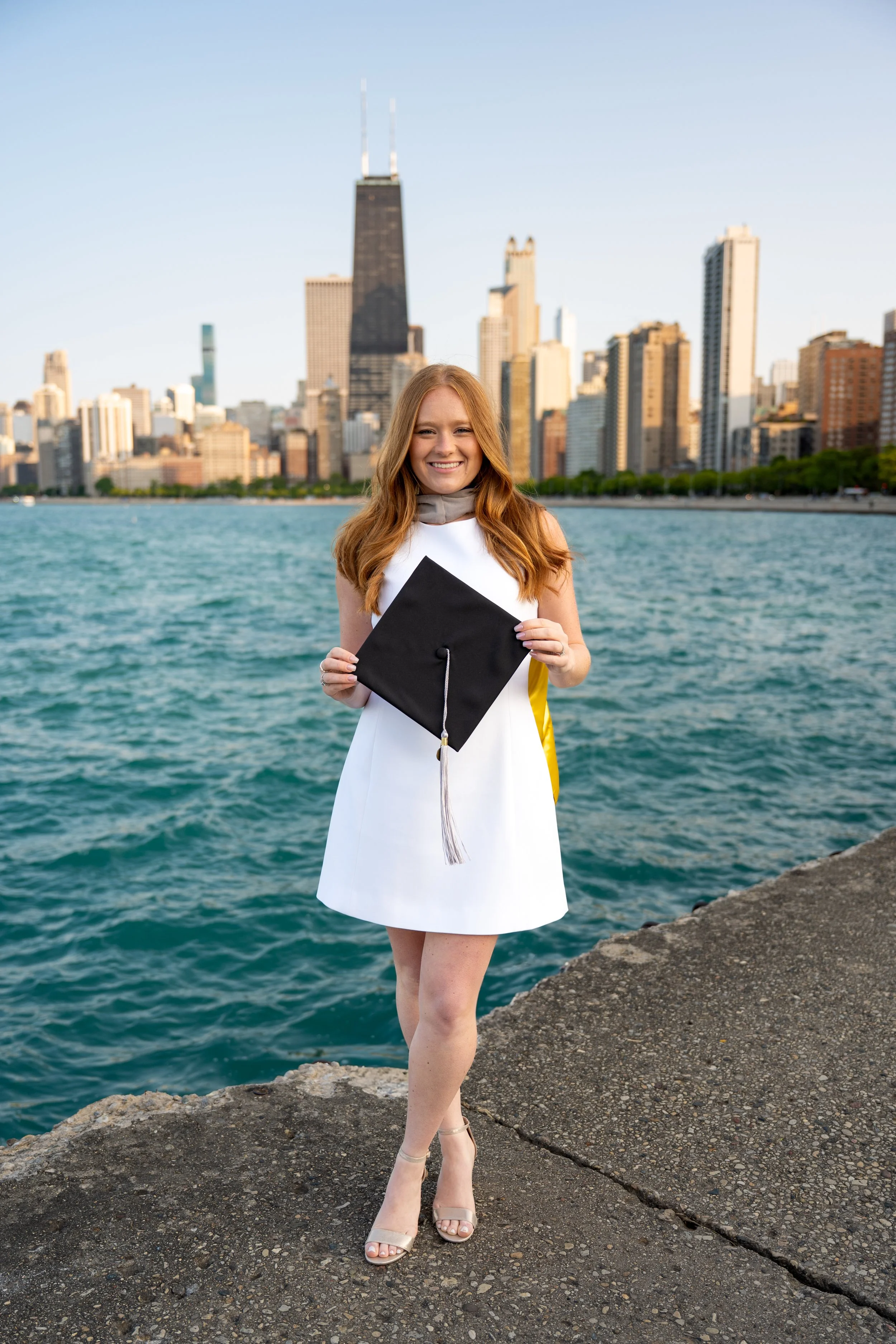A young woman with long red hair smiling while holding a graduation cap, standing on rocks near a body of water with a city skyline, including the John Hancock Center and Willis Tower, in the background.