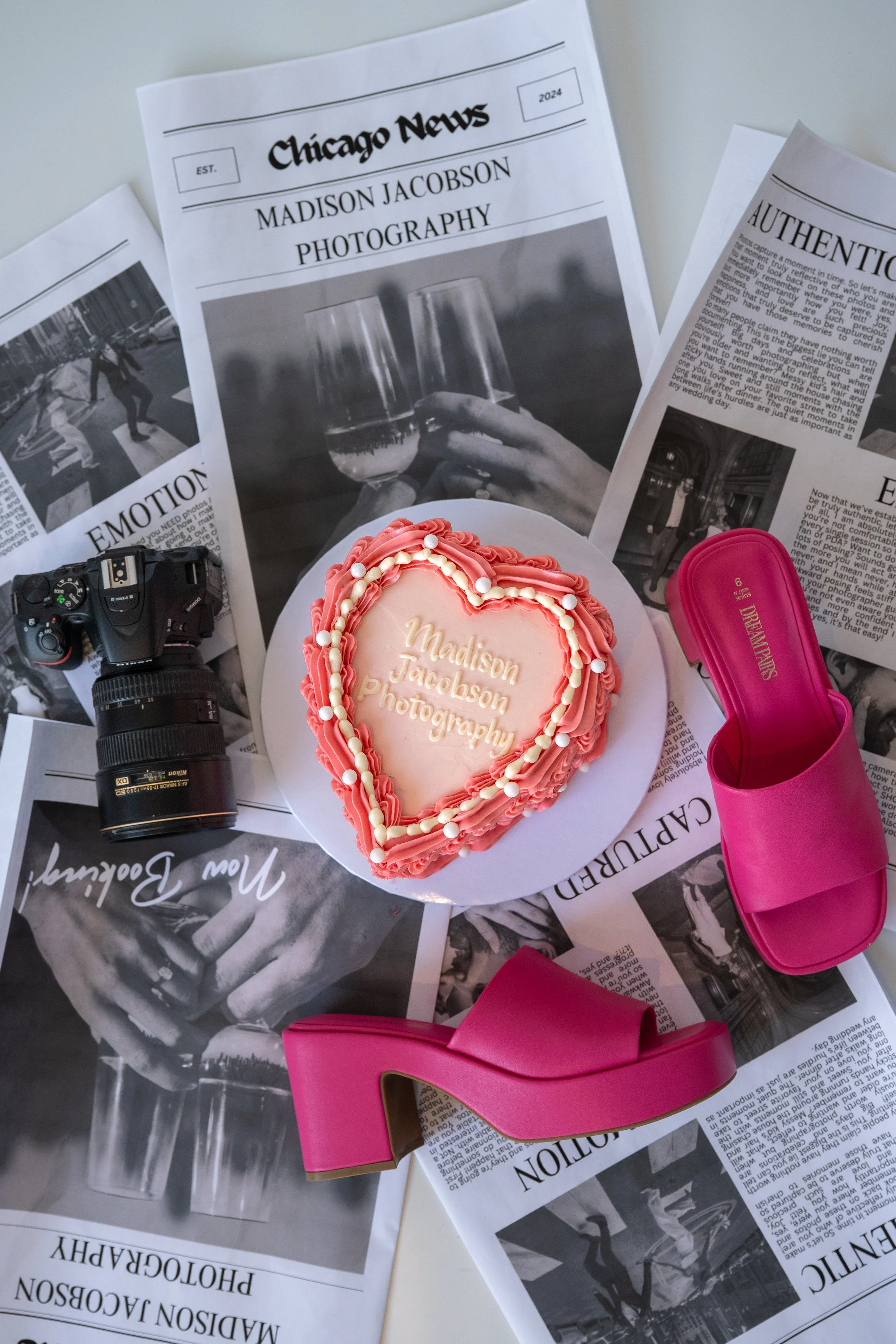 A pink heart-shaped cake with white and pink icing, inscribed with 'Madison Jacobson Photography.' Surrounding it are black and white newspapers, a camera, and pink high-heeled sandals.