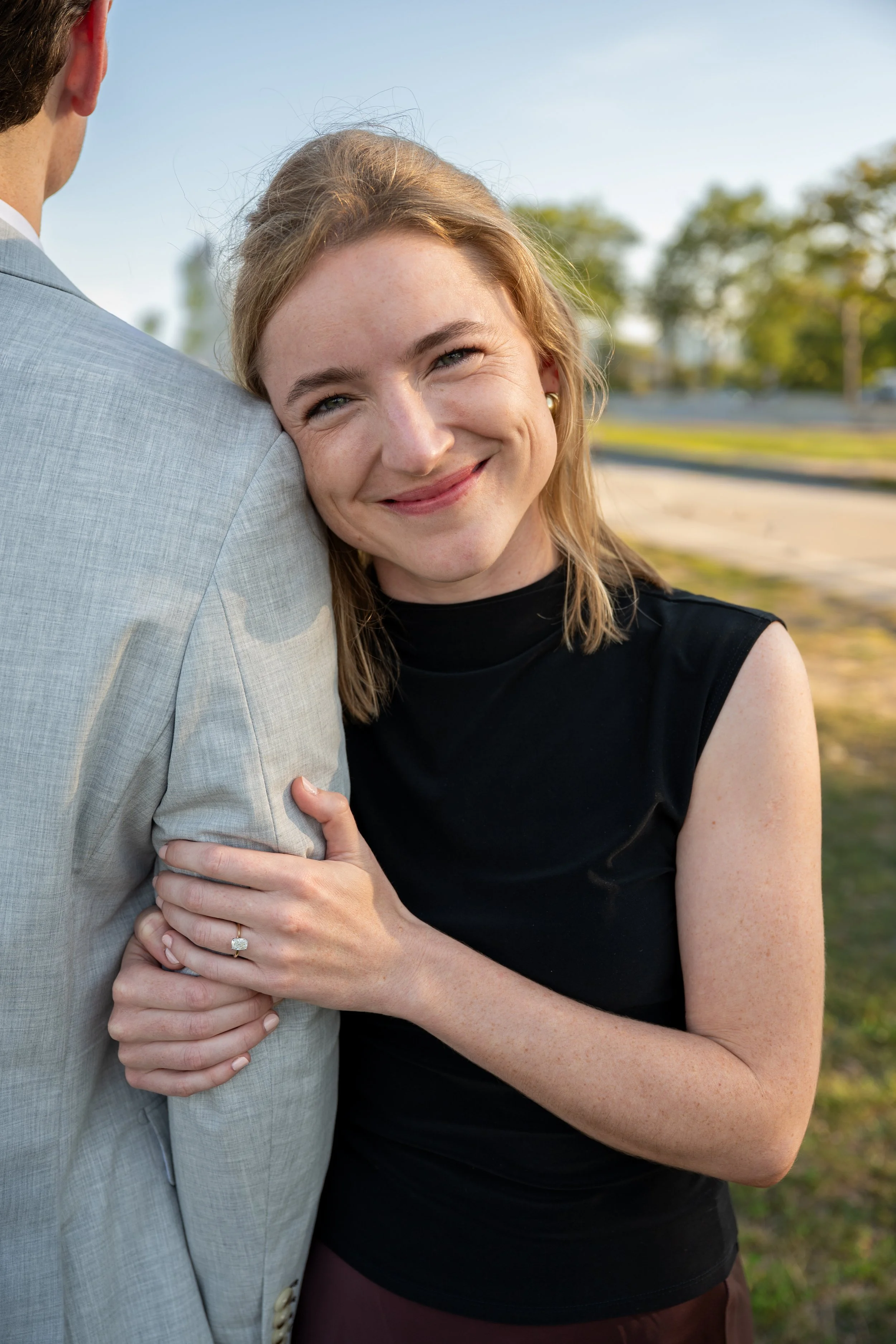 A woman with blonde hair smiling and hugging a man whose face is not visible. She is wearing a black sleeveless top and has a diamond ring on her finger. They are outdoors with trees and a partly cloudy sky in the background.