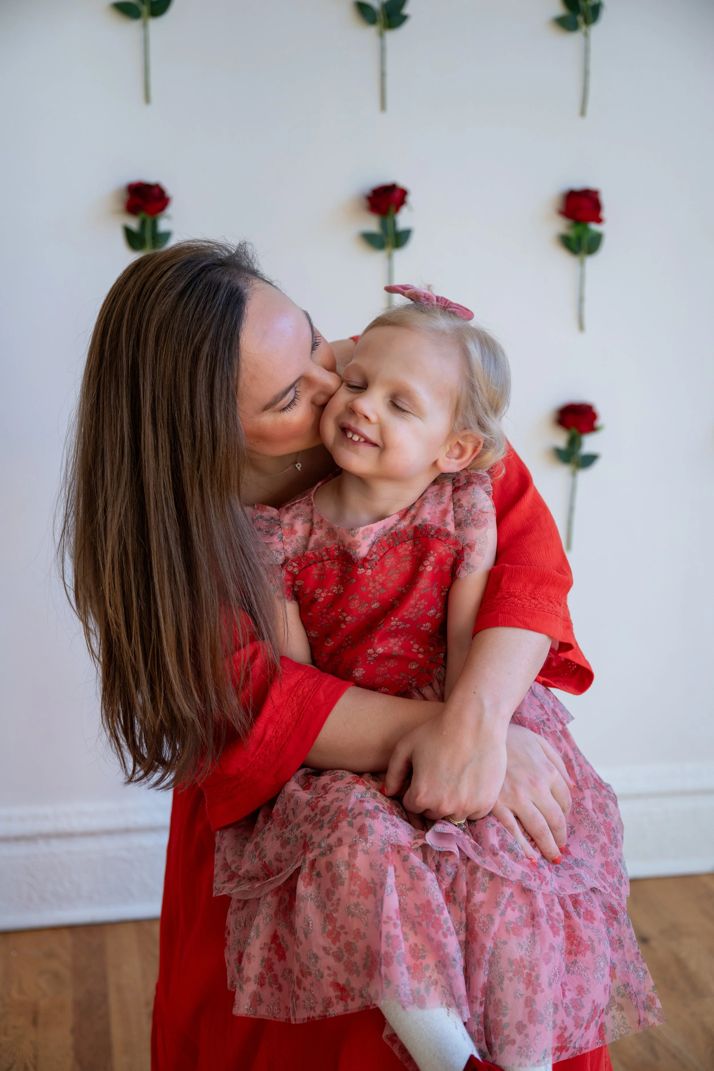 A woman with long brown hair is holding a young girl with blonde hair, both smiling and sharing a kiss on the cheek. The woman is wearing a red dress, and the girl is dressed in a pink floral dress with white tights. In the background, a white wall is decorated with red roses and green leaves.
