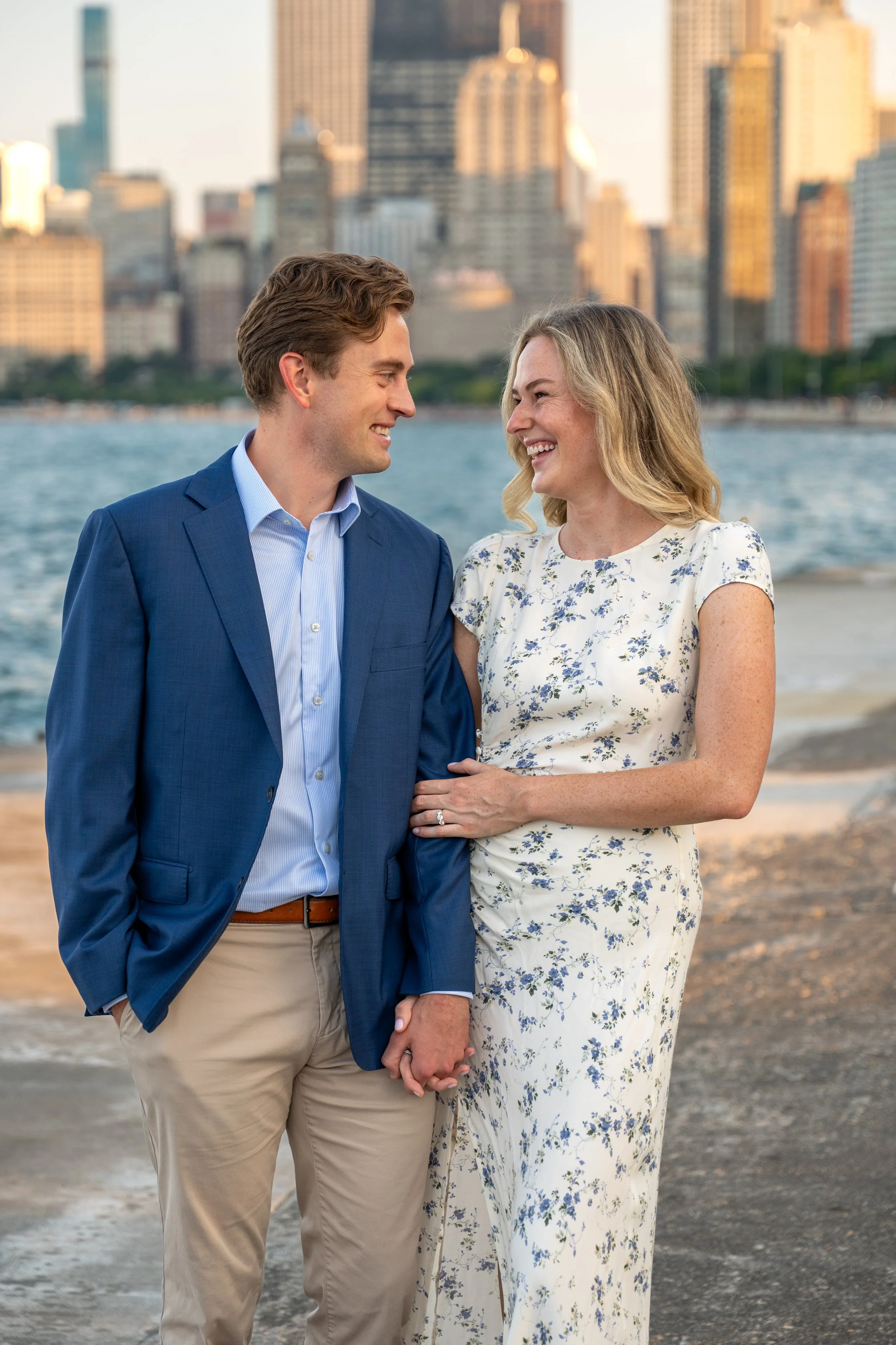 A couple smiling and holding hands near the water with the Chicago skyline in the background.