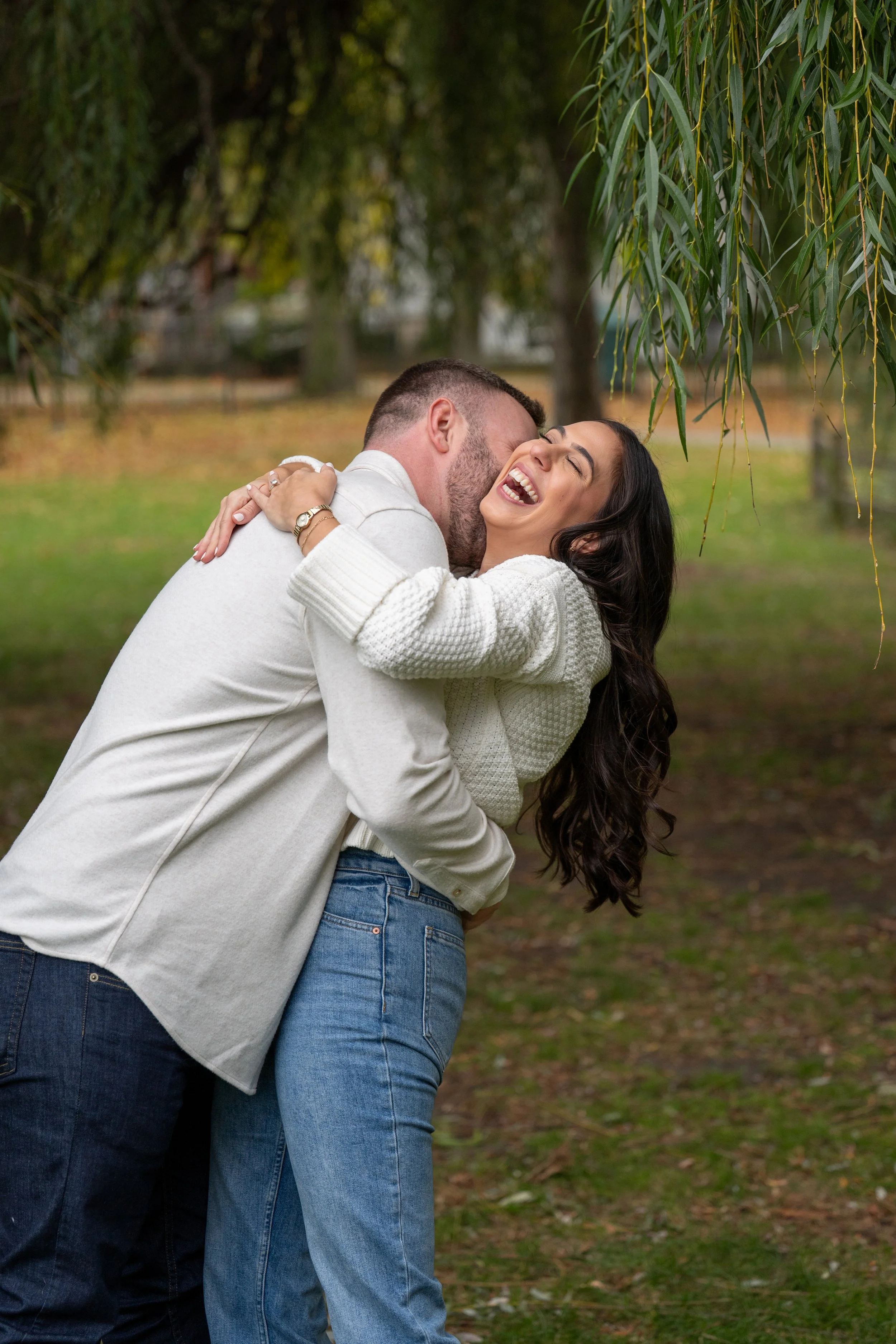 Photography of a couple hugging and laughing outdoors in a park with trees and green grass, during daytime.