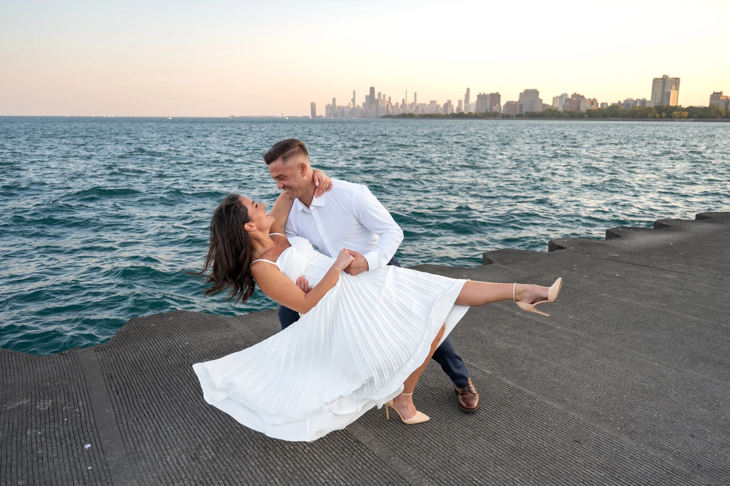 A couple by the water, the man lifting the woman as she lies in his arms, with the Chicago city skyline in the background. The woman is wearing white heels and a flowing white, Marilyn Monroe style dress.