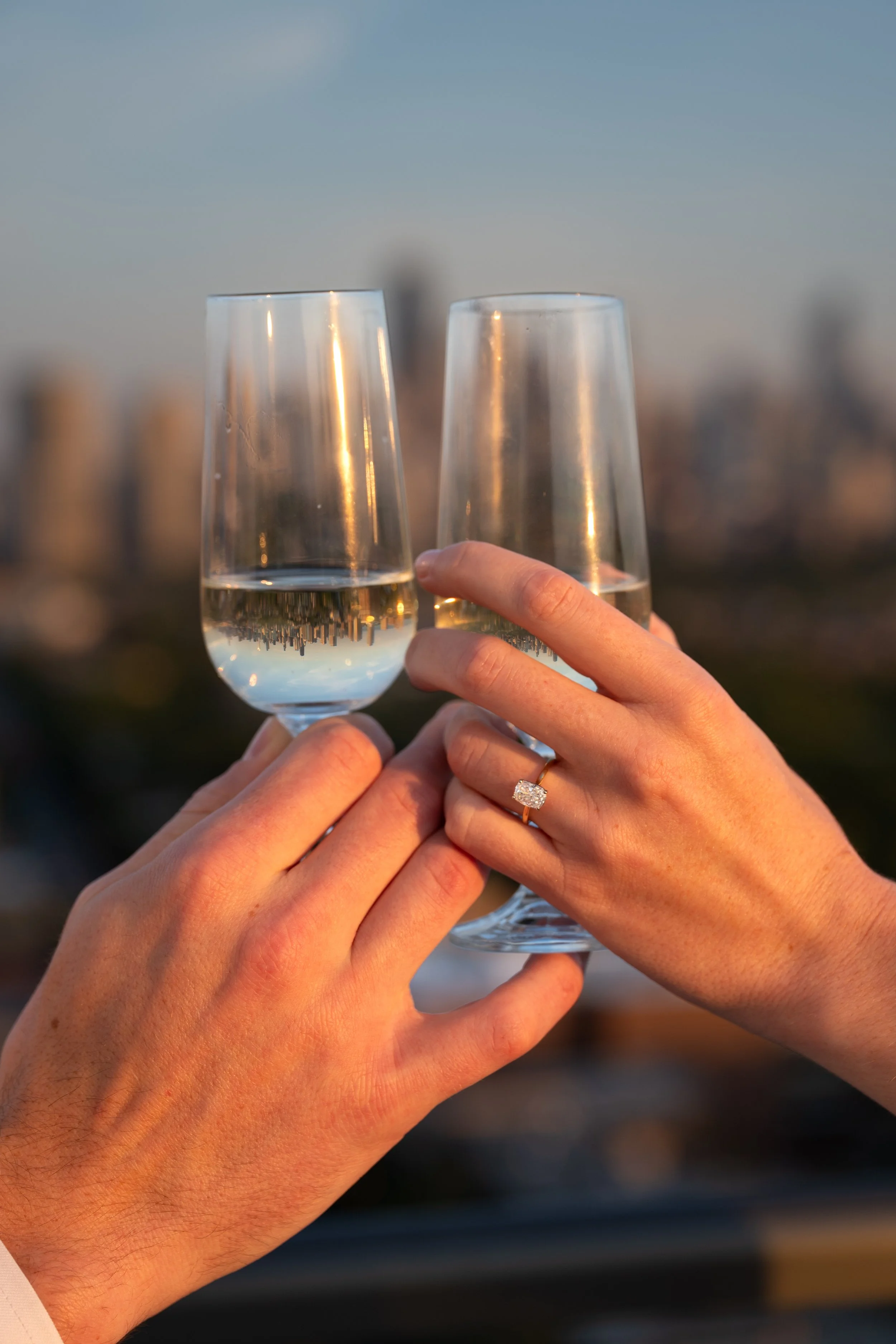 Two hands holding champagne glasses with the Chicago skyline in background at sunset. The woman is wearing an engagement ring.