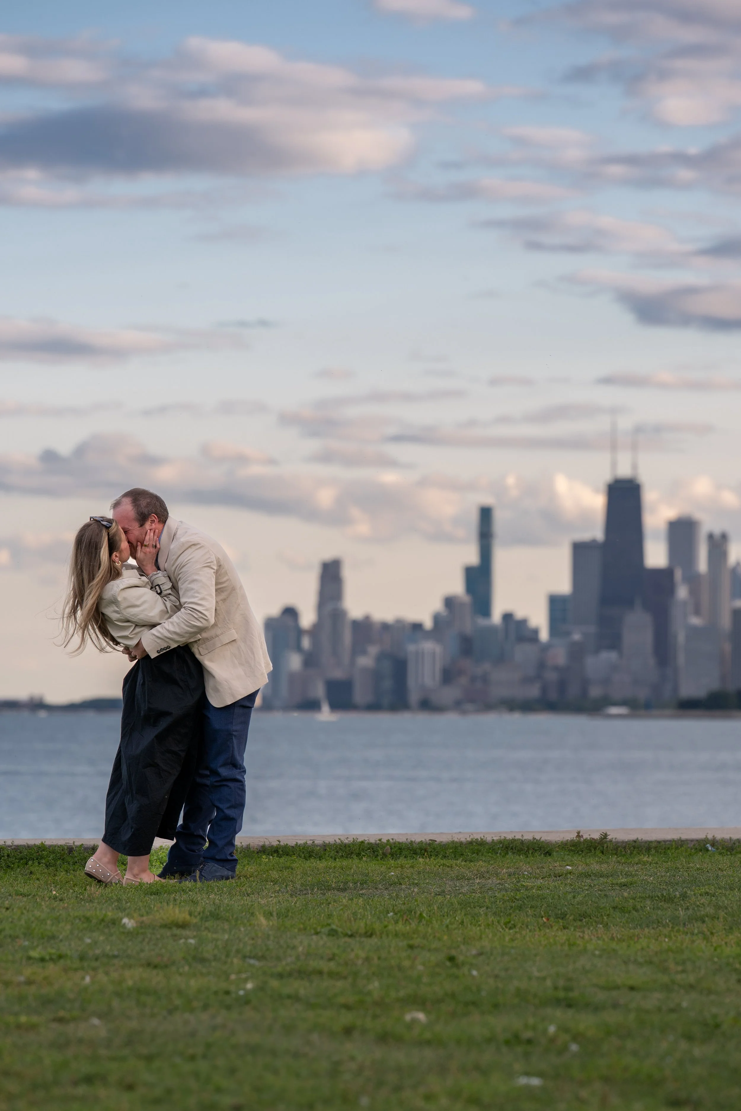 A couple kissing outdoors by a body of water during sunset with the Chicago city skyline in the background.
