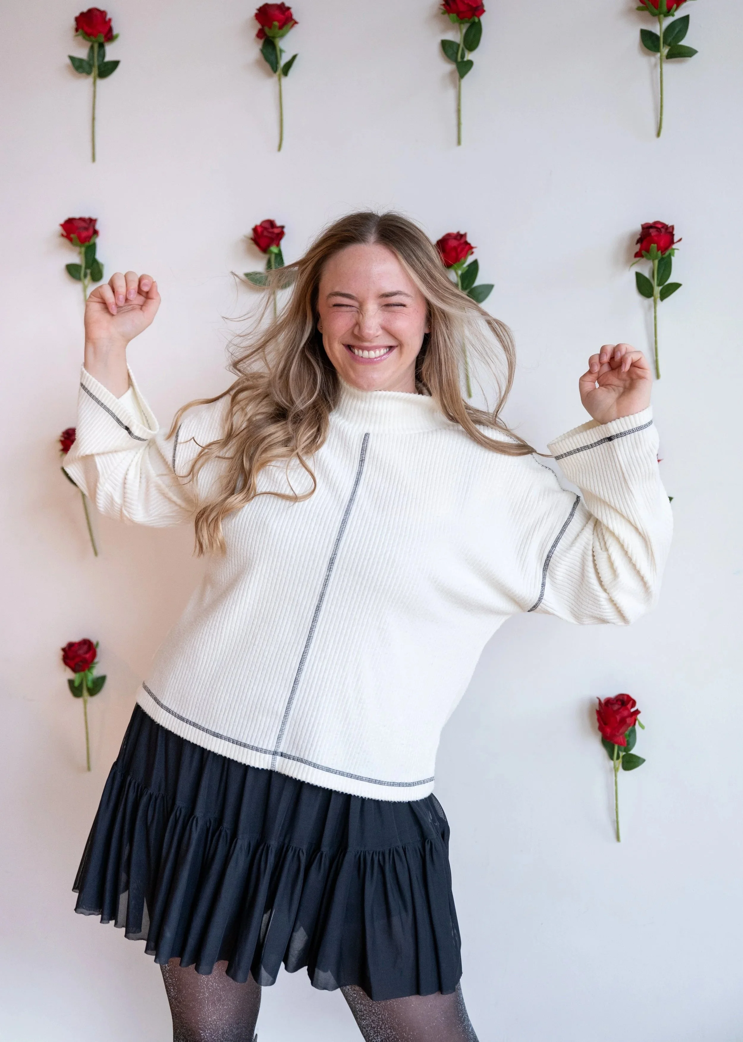 A woman with long blonde hair smiling and dancing in front of a white wall decorated with red roses. She is wearing a white long-sleeve jacket, a flowy black miniskirt, and lace black leggings.