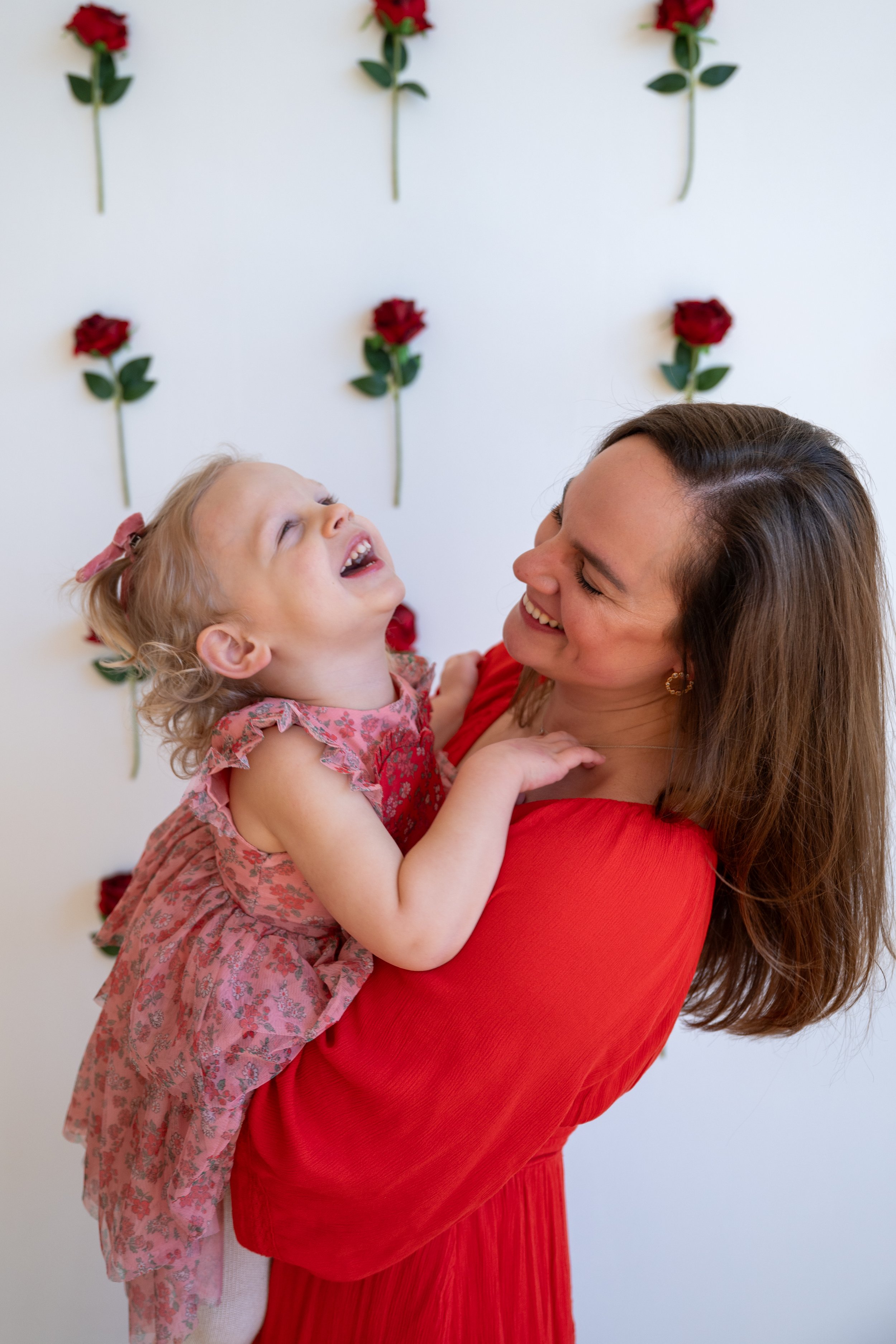 A woman with long brown hair in a red dress holding a little girl with curly blonde hair, wearing a pink floral dress, against a white wall with red roses.