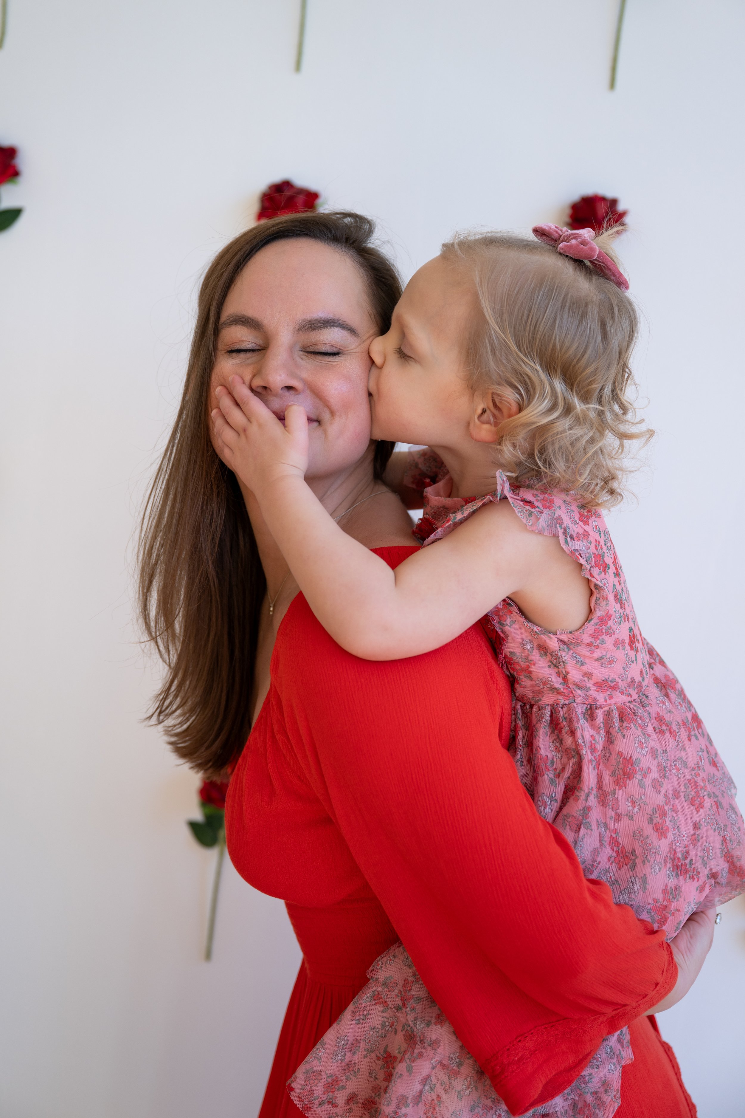 A woman with long brown hair smiling as a young girl with curly blonde hair kisses her cheek. The girl is wearing a pink floral dress and pink bow in her hair, and the woman is wearing a red dress. They are indoors with a white wall decorated with red roses and green leaves in the background.