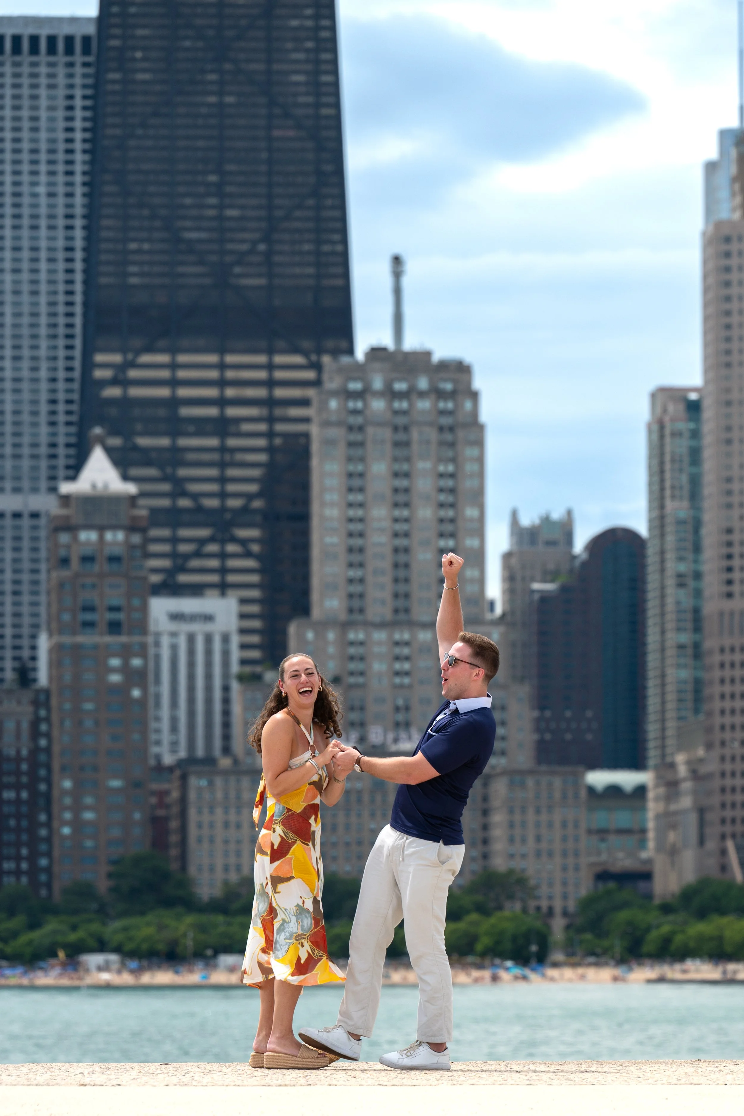 Photography of a man and woman standing on a beach, dressed in formal attire, holding hands. The man is celebrating that he just proposed and there are Chicago skyscrapers in the background.