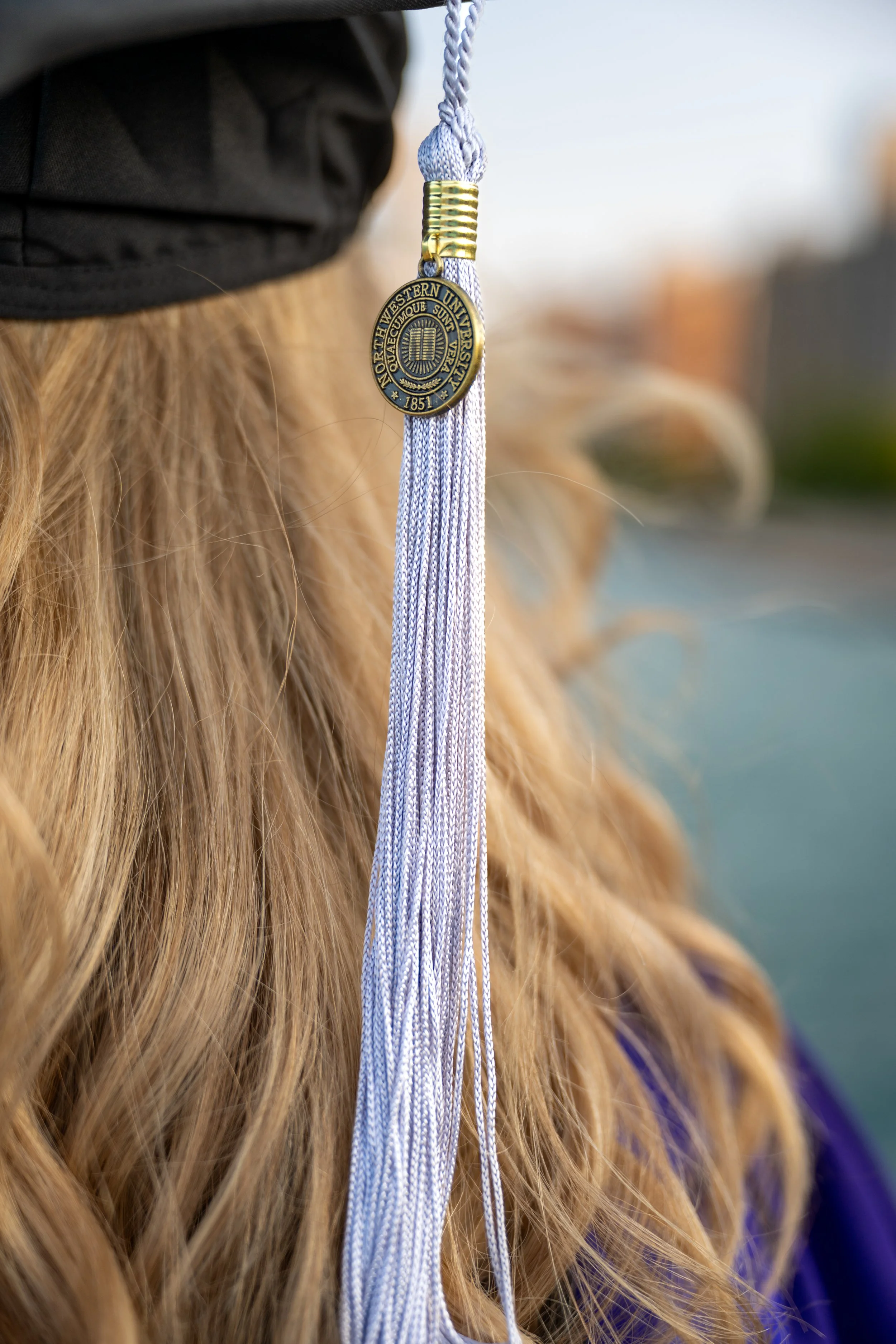A close-up of a college graduation cap's tassel hanging on a student's head, with long, wavy hair visible, and a blurred background.