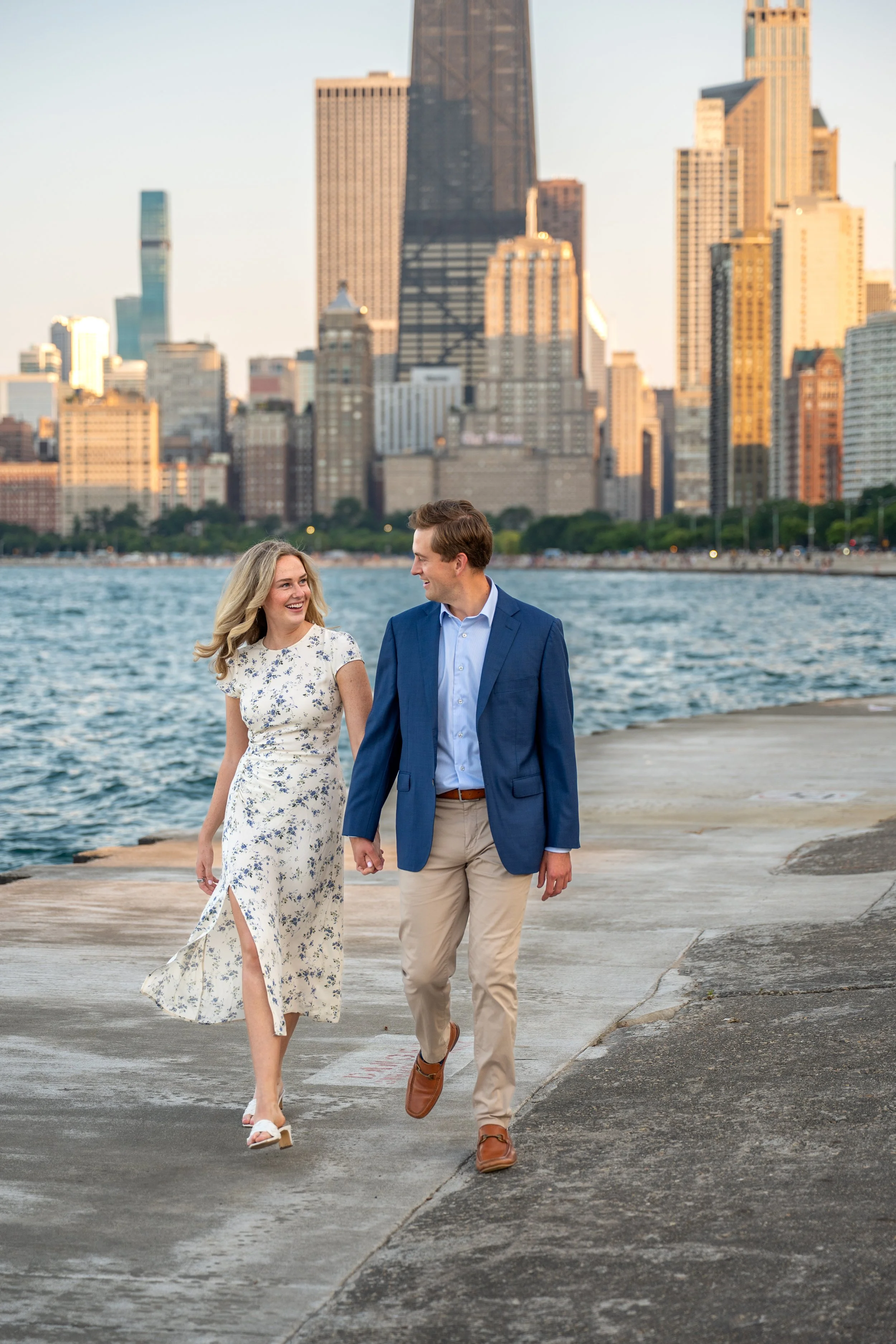 Photography of a woman and a man walking hand in hand along a waterfront walkway with the Chicago skyline in the background, smiling at each other.