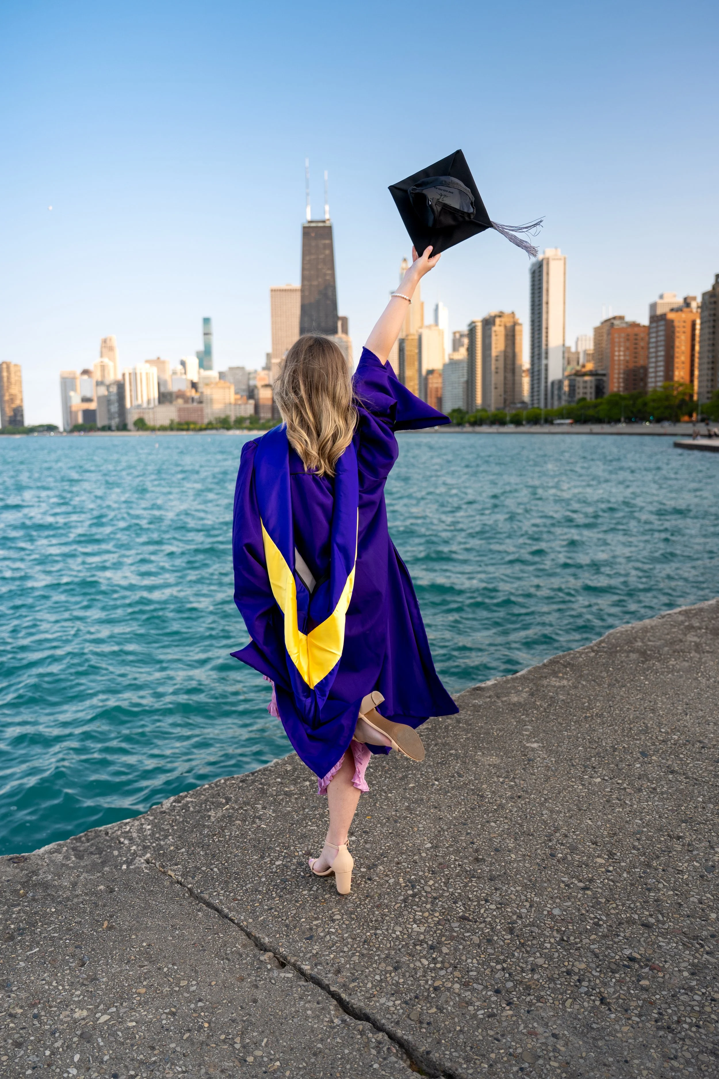 A woman in a purple graduation gown and heels is standing on a stone platform near a body of water, holding her graduation cap in the air. The city skyline, including tall skyscrapers and Lake Michigan, is in the background during sunset.