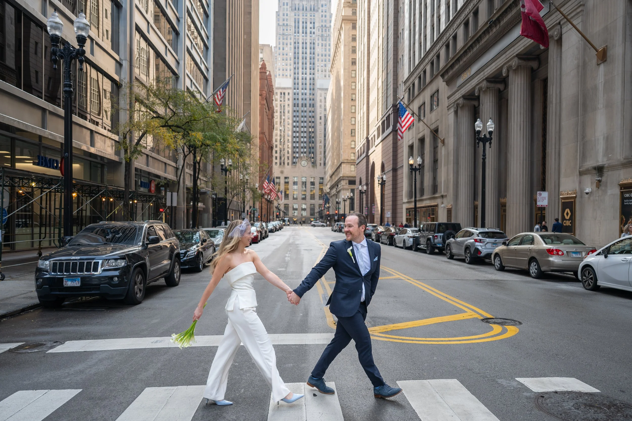 A bride and groom in wedding attire holding hands and walking across a Chicago city street at a pedestrian crossing. Tall buildings line the street, which is decorated with flags and street lamps.