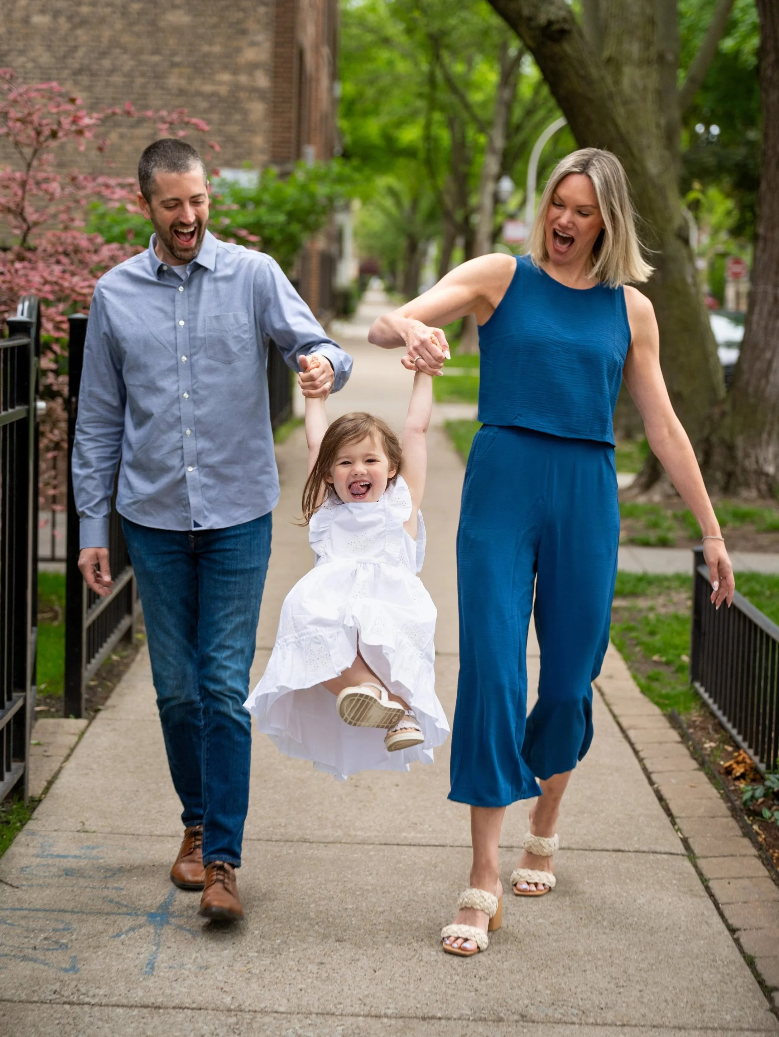 A family of three walking on a sidewalk in a neighborhood with trees and brick houses. The adults are smiling and holding a young girl between them, who is smiling and looking happy.