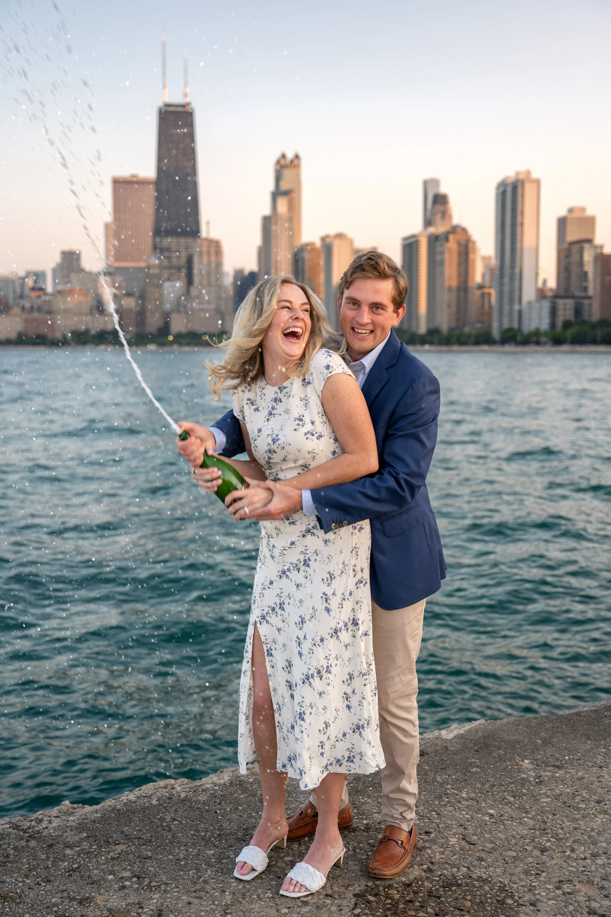 A happy couple is by the water, with the Chicago skyline in the background, celebrating with a spraying champagne bottle.