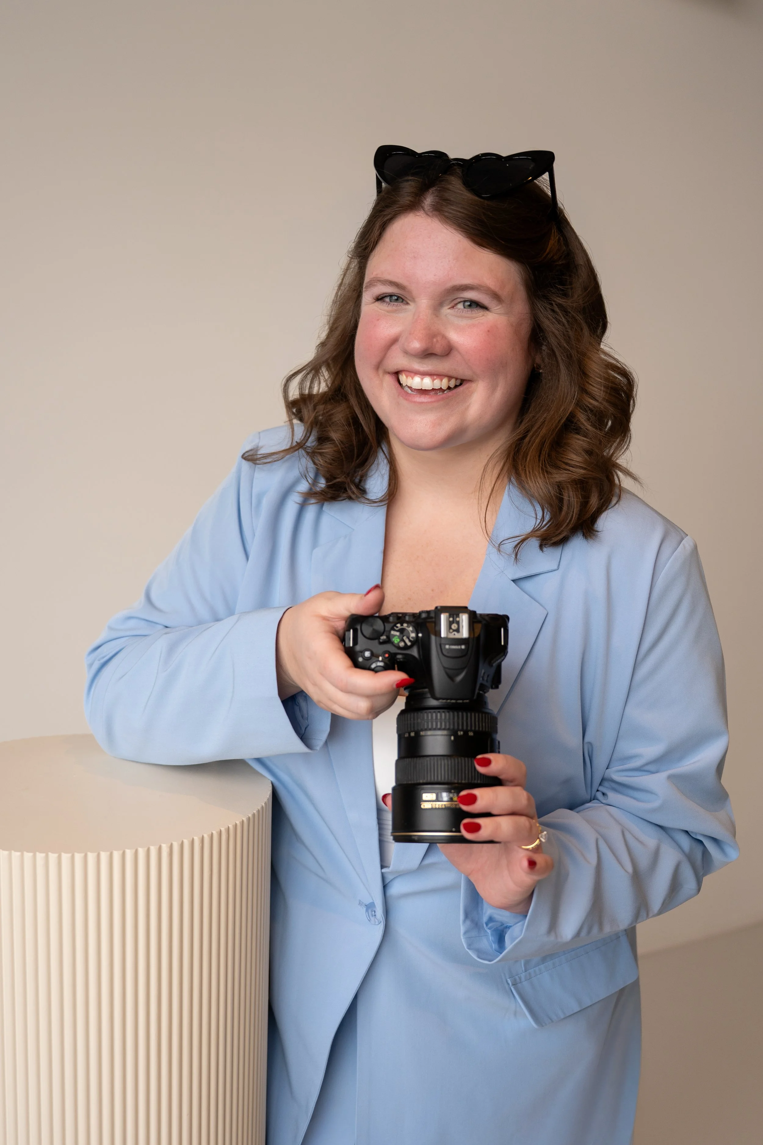 A smiling female photographer with brown hair wearing a light blue blazer, black heart-shaped sunglasses, and is holding a camera with a large zoom lens.