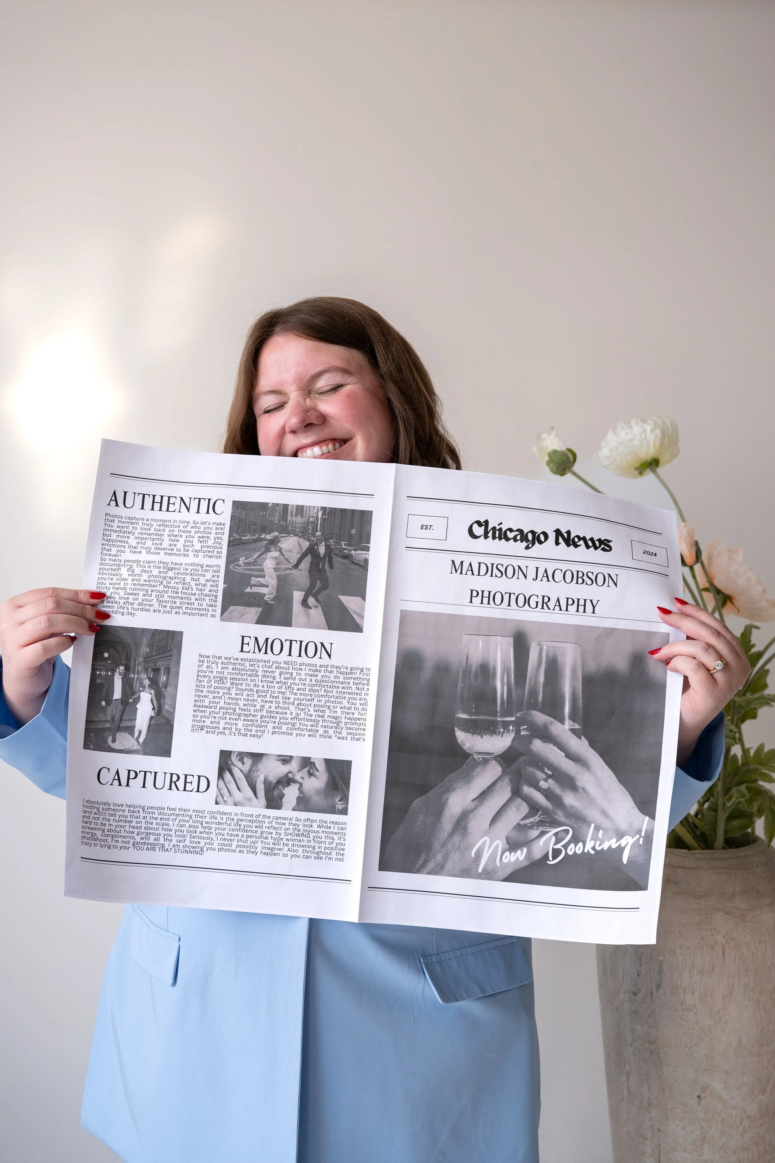 A woman with long brown hair smiling and holding a large newspaper that says Chicago News: Madison Jacobson Photography. She is wearing a light blue blazer and has red nail polish. In the background is a vase of white flowers.