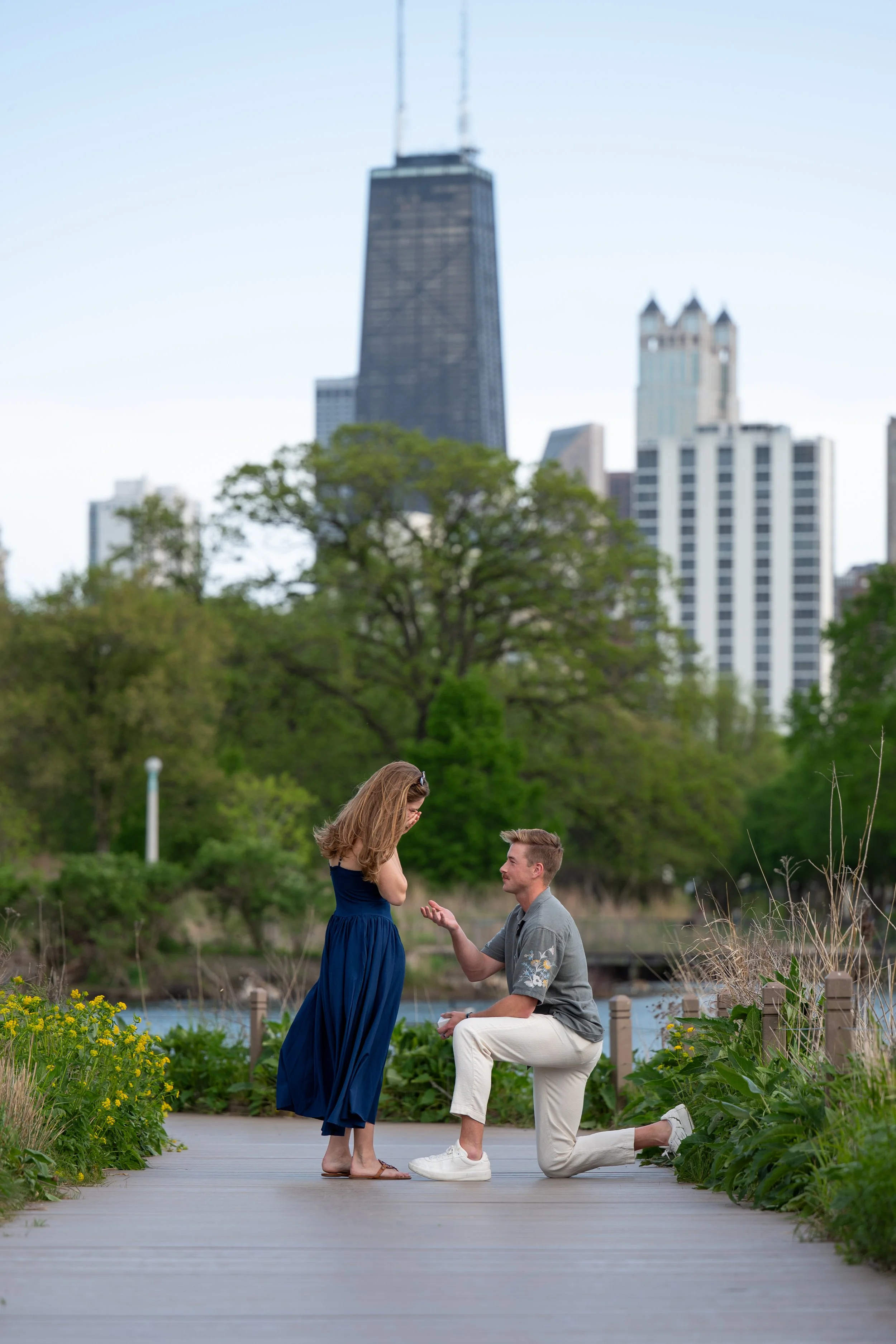 A man proposing marriage to a woman outdoors on a wooden pathway, with Chicago city skyscrapers in the background and lush green trees surrounding them.