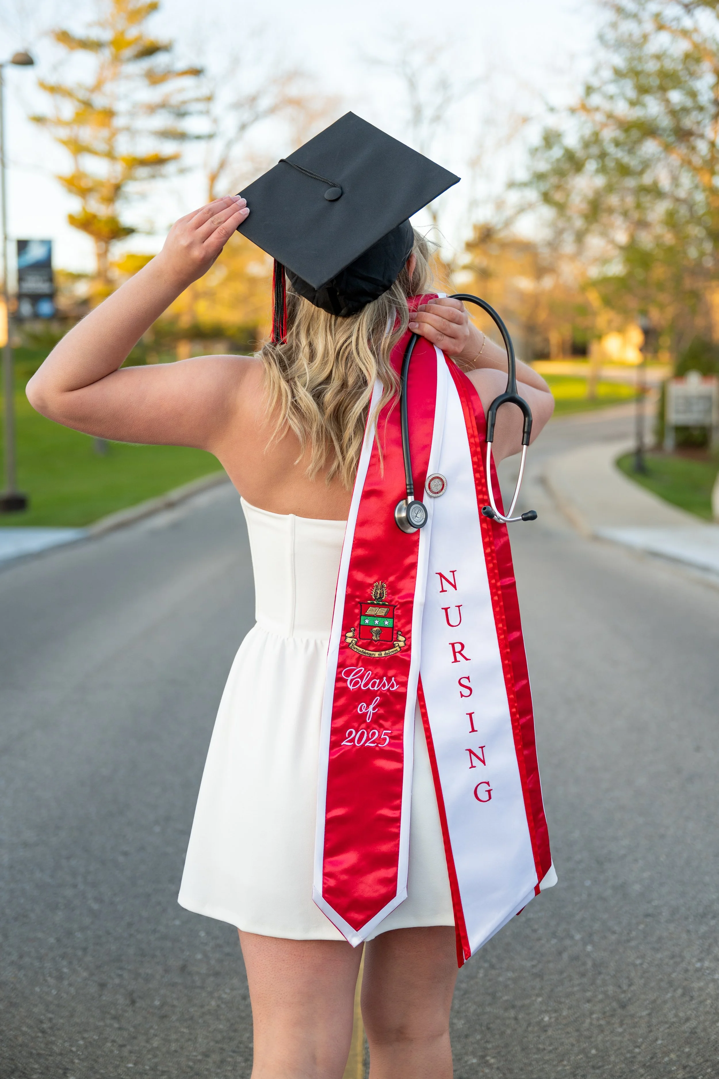 A woman in a white dress, graduation cap, and red and white sash holding a stethoscope over her shoulder during graduation, standing on an empty street with trees in the background.