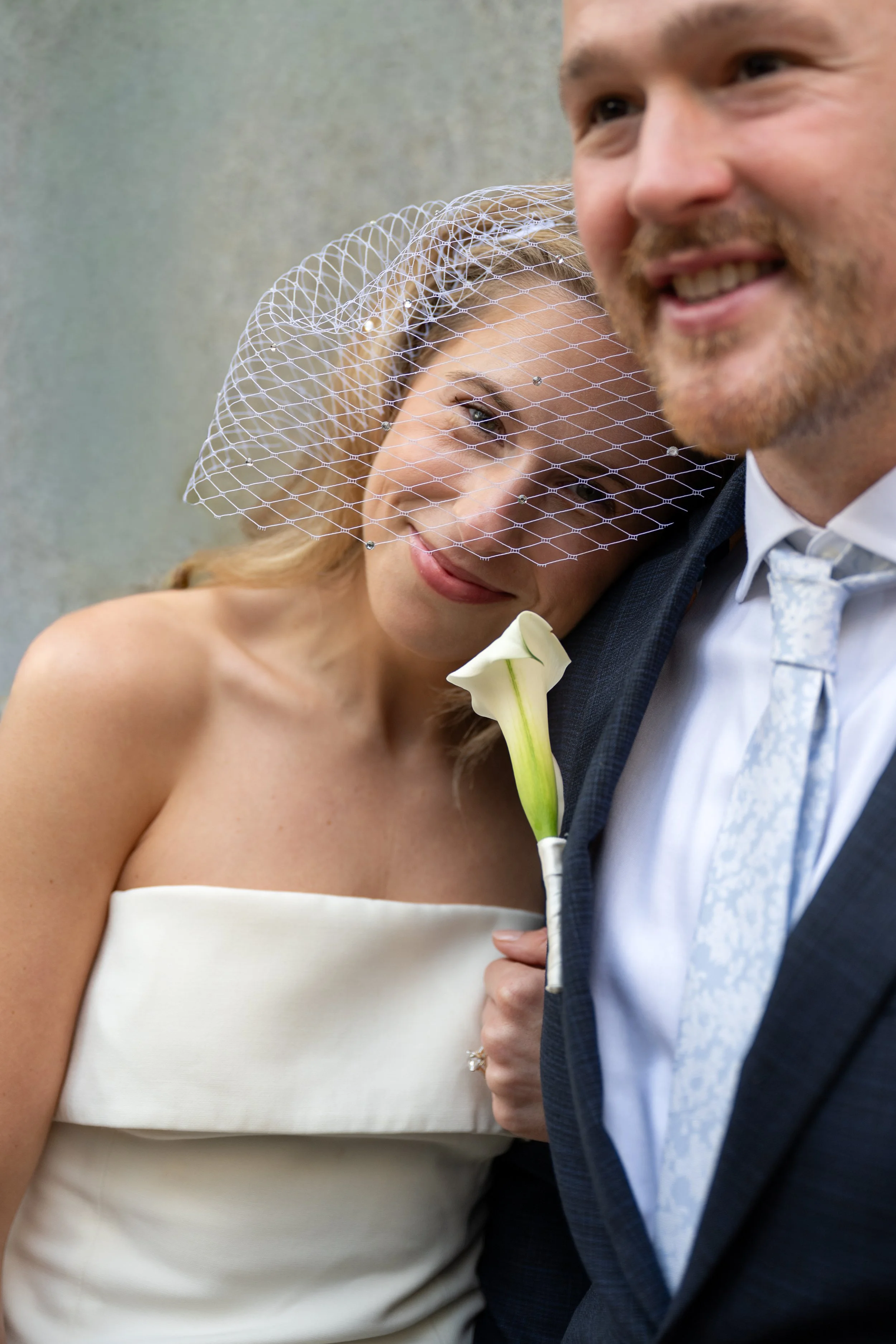 A newly married couple smiling and close together, with the woman resting her head on the man's shoulder. The woman is wearing a white birdcage veil, white top, and holding a calla lily. The man is dressed in a suit with a light-colored tie.