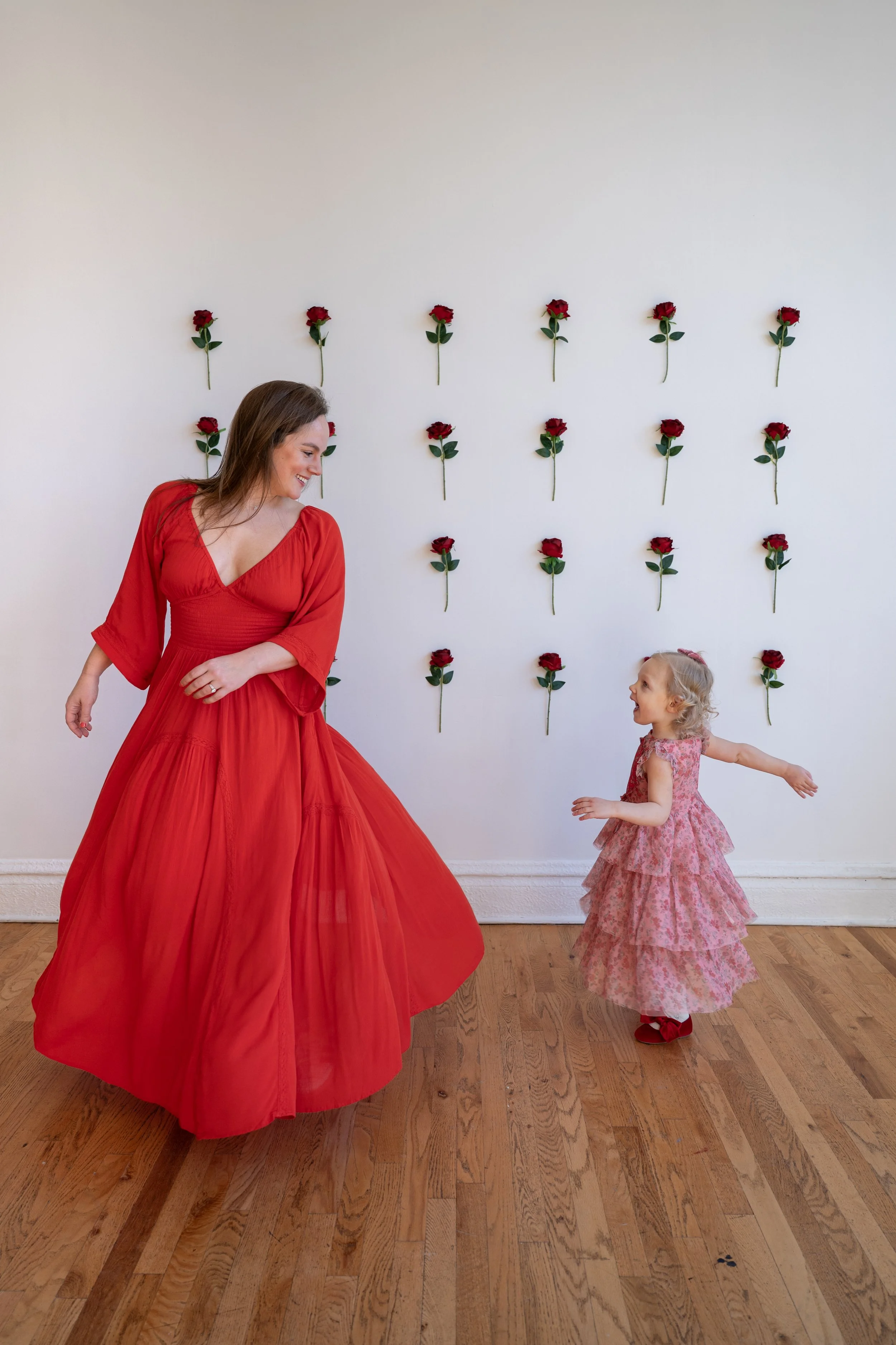 A woman and a girl dancing together indoors against a white wall decorated with red roses. The woman is wearing a long, flowing red dress, and the girl is wearing a pink floral dress.