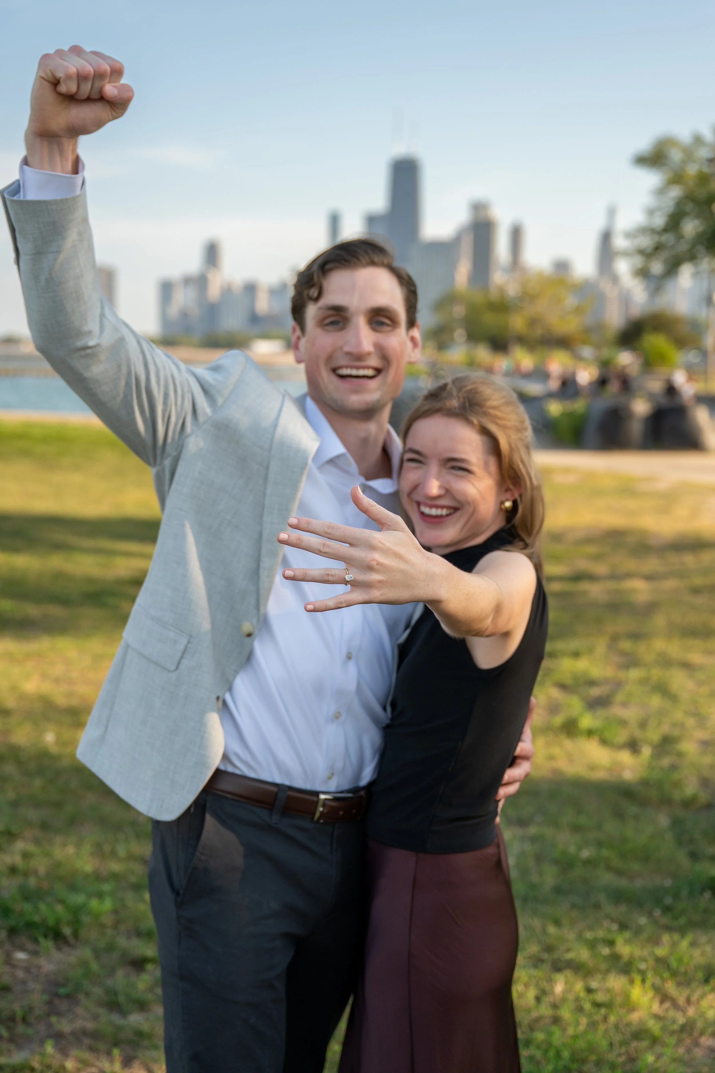 A happy couple in a park with the Chicago city skyline in the background, celebrating with the man raising his fist and the woman showing her engagement ring.