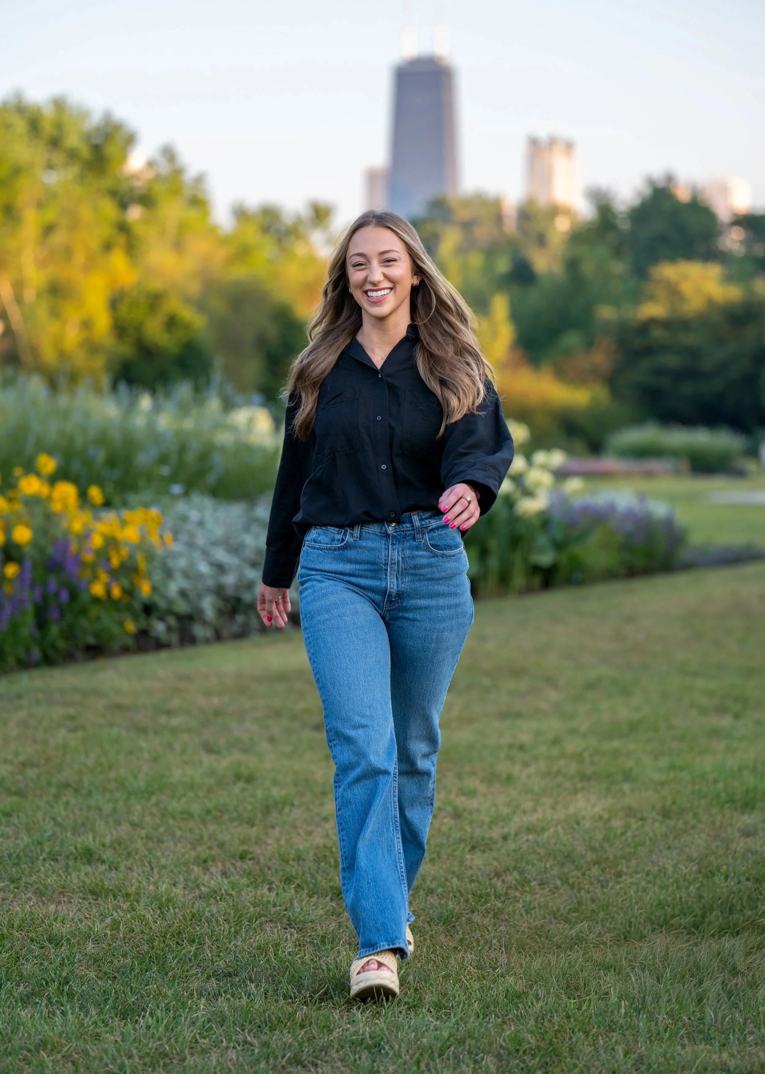 Photography of a smiling young woman with long wavy hair walking in a park with green grass, colorful flowers, and trees in the background, with the Chicago skyline in the distance.
