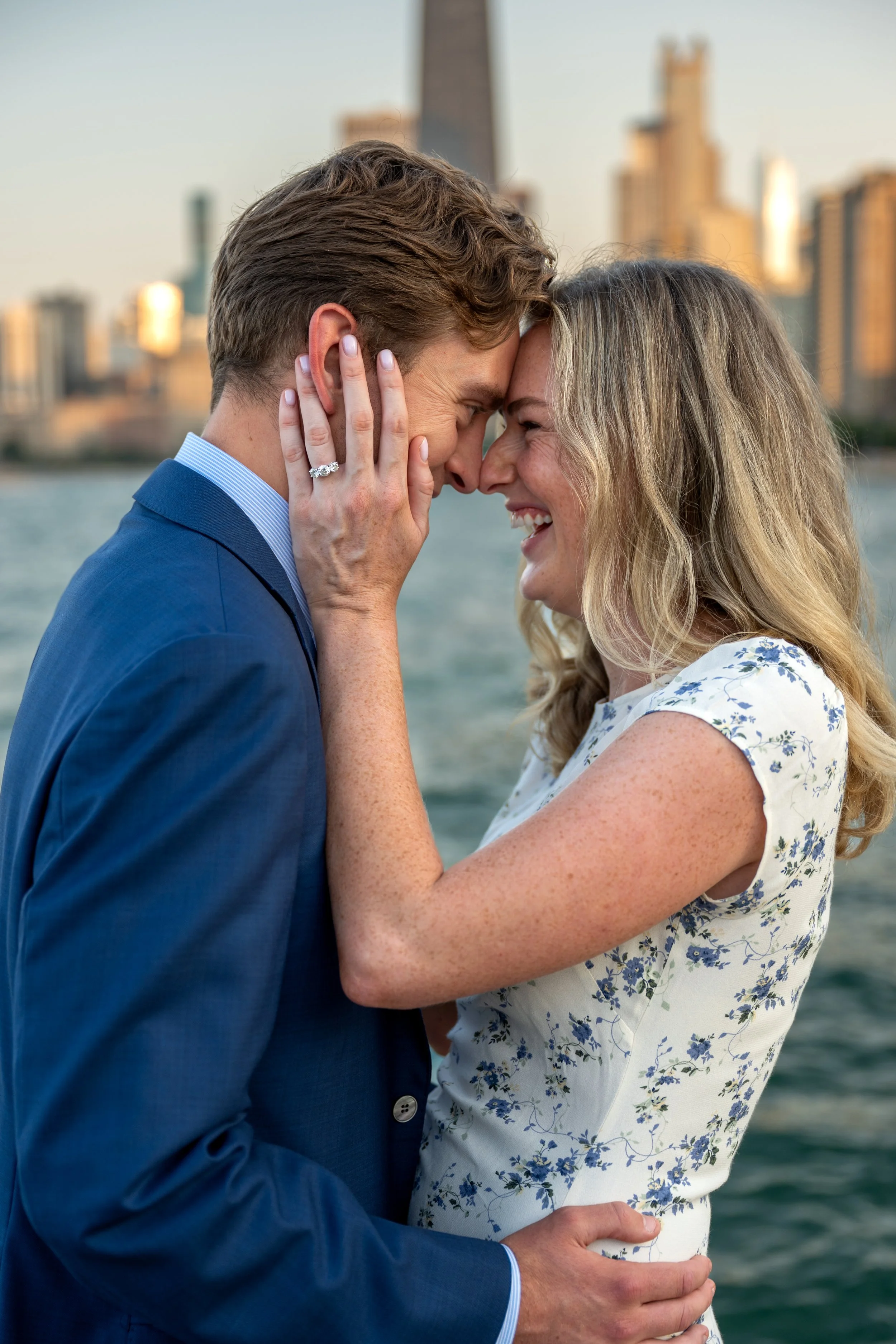 A couple in formal attire embracing by a body of water with the Chicago skyline in the background, touching foreheads and smiling.