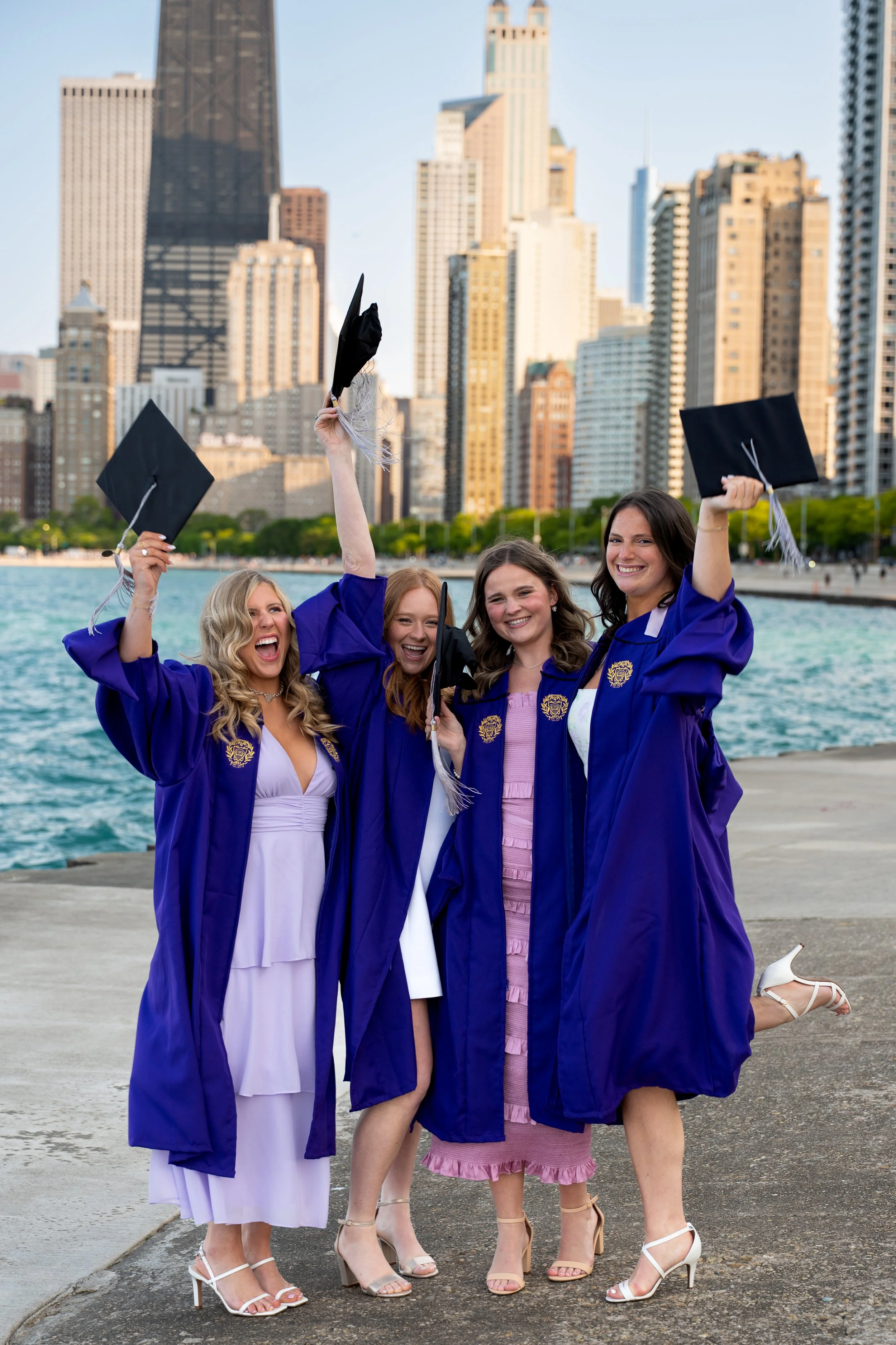 Four women in purple graduation gowns celebrating by the water with the Chicago city skyline in the background, holding graduation caps.