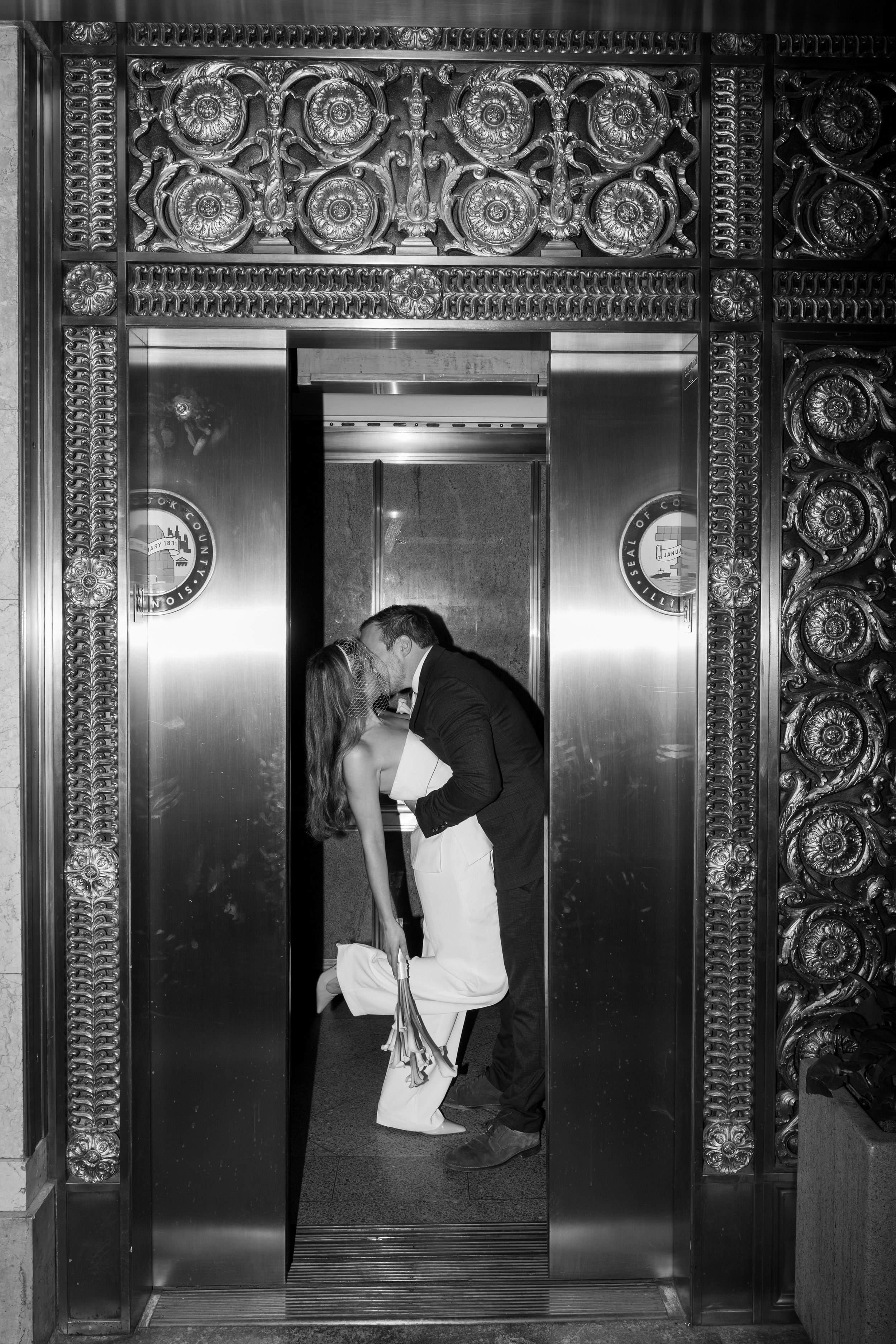 A couple kissing in front of an ornate elevator door. The woman is dressed in a white jumpsuit, and the man is wearing a suit. The elevator door has intricate decorative carvings.