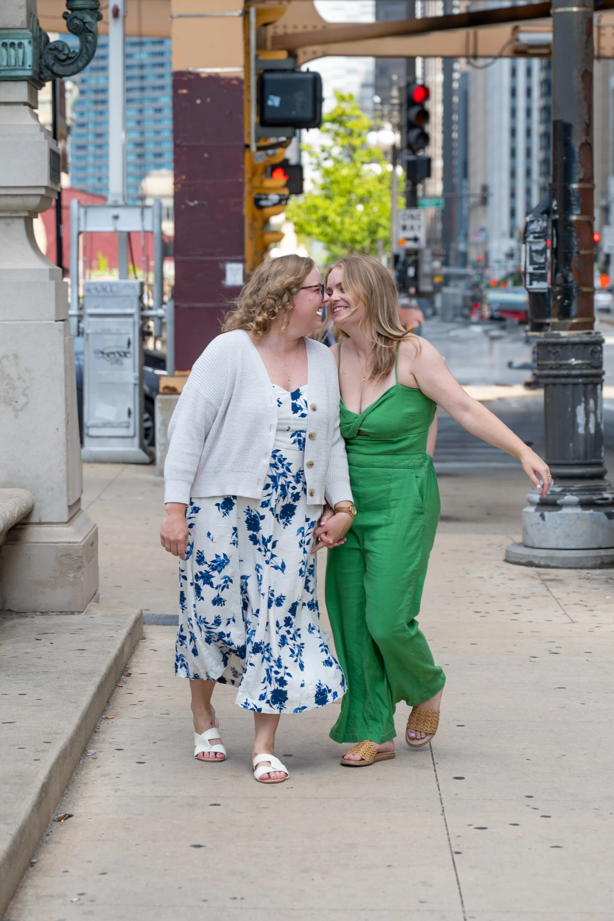 Two women holding hands and smiling as they walk down a Chicago city street with traffic signals and tall buildings in the background.
