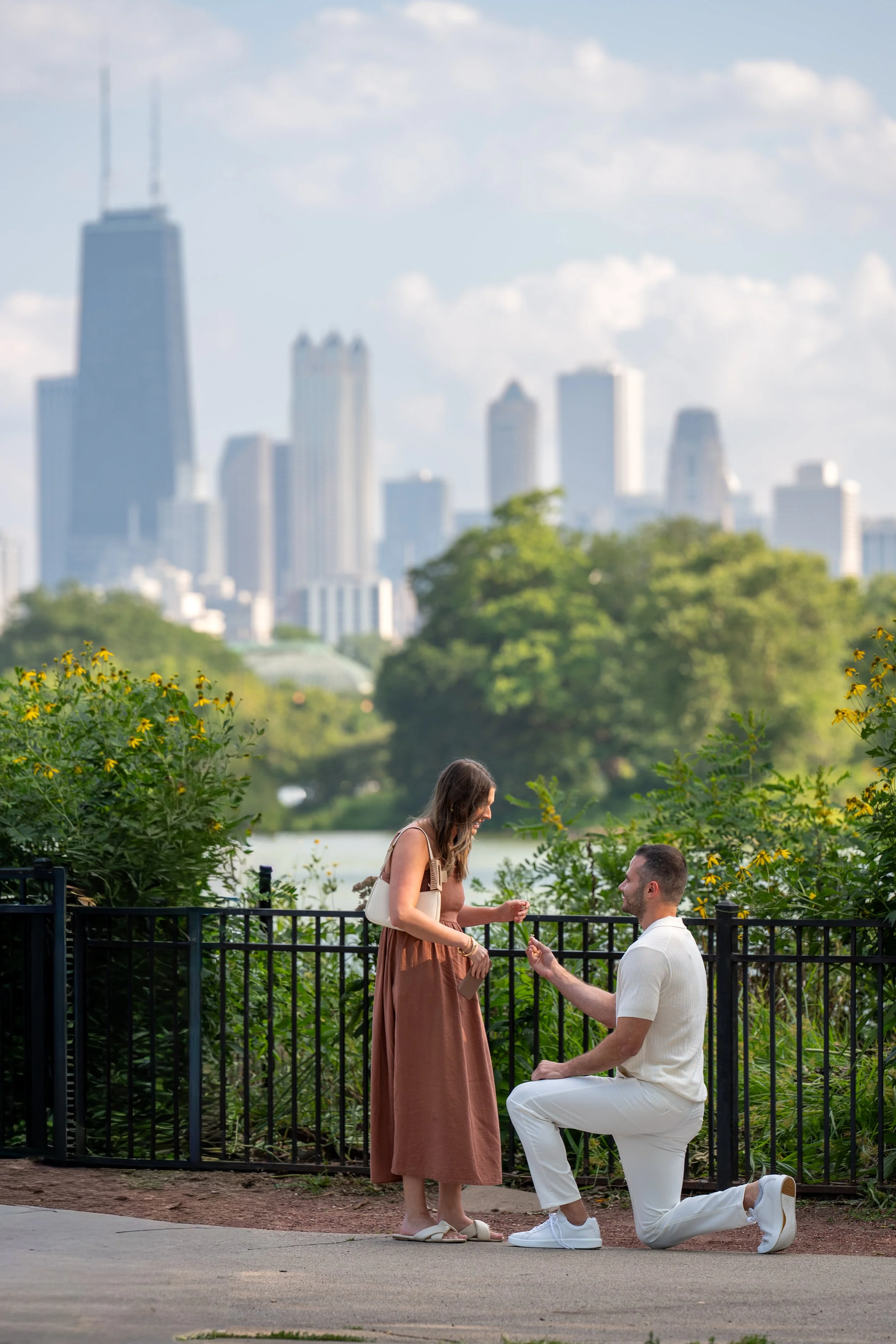A man proposing marriage to a woman in a park with the Chicago city skyline in the background during daytime.