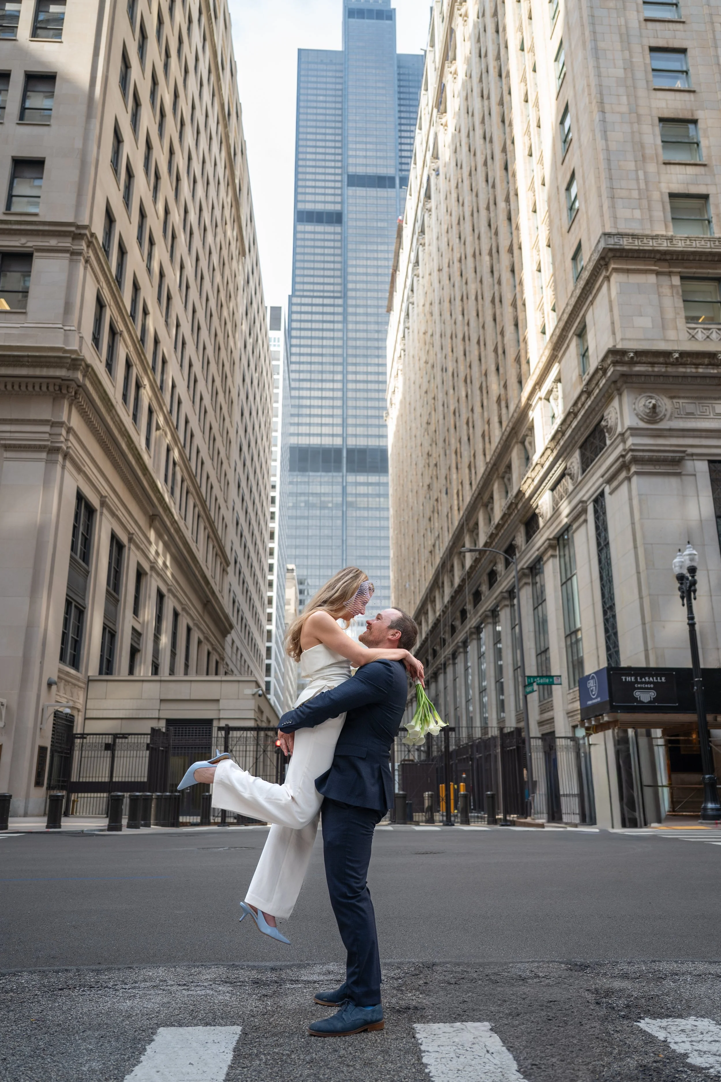 A man in a navy suit lifting a woman in a white jumpsuit in the air on a city street in Chicago, with tall buildings in the background, during daytime.