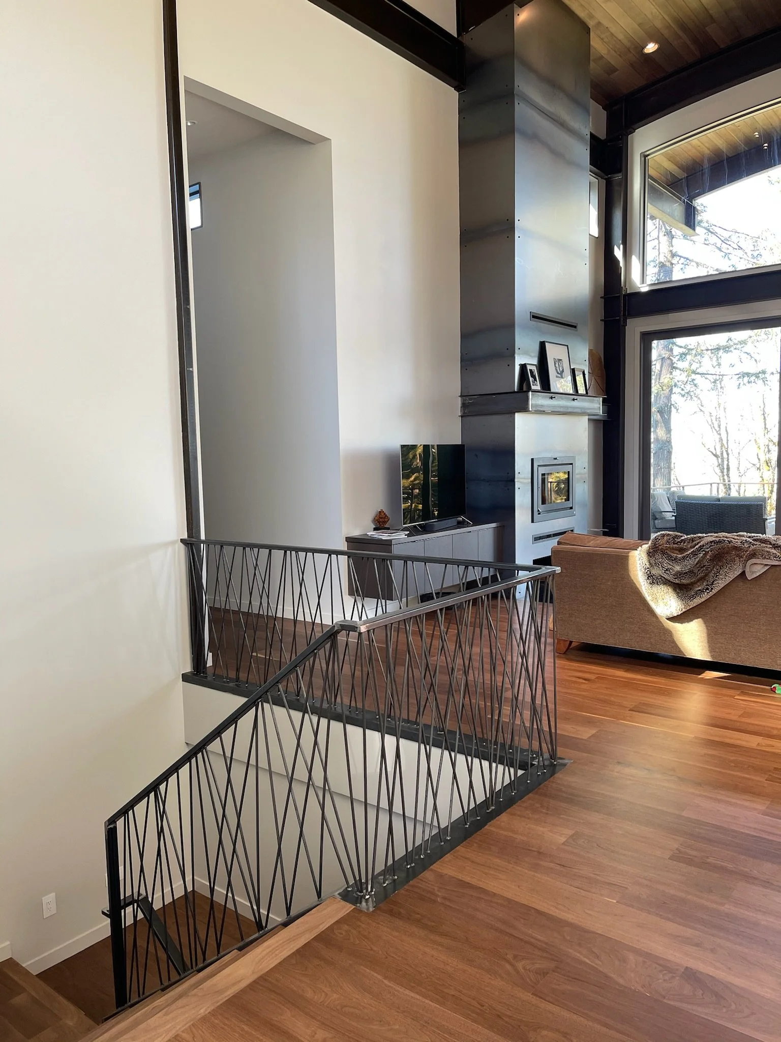 Interior view of a modern living room with a staircase featuring black metal railing, a fireplace with a tall metallic chimney, a TV on a stand, framed photos, and a beige sofa near large windows showing a view of trees outside.
